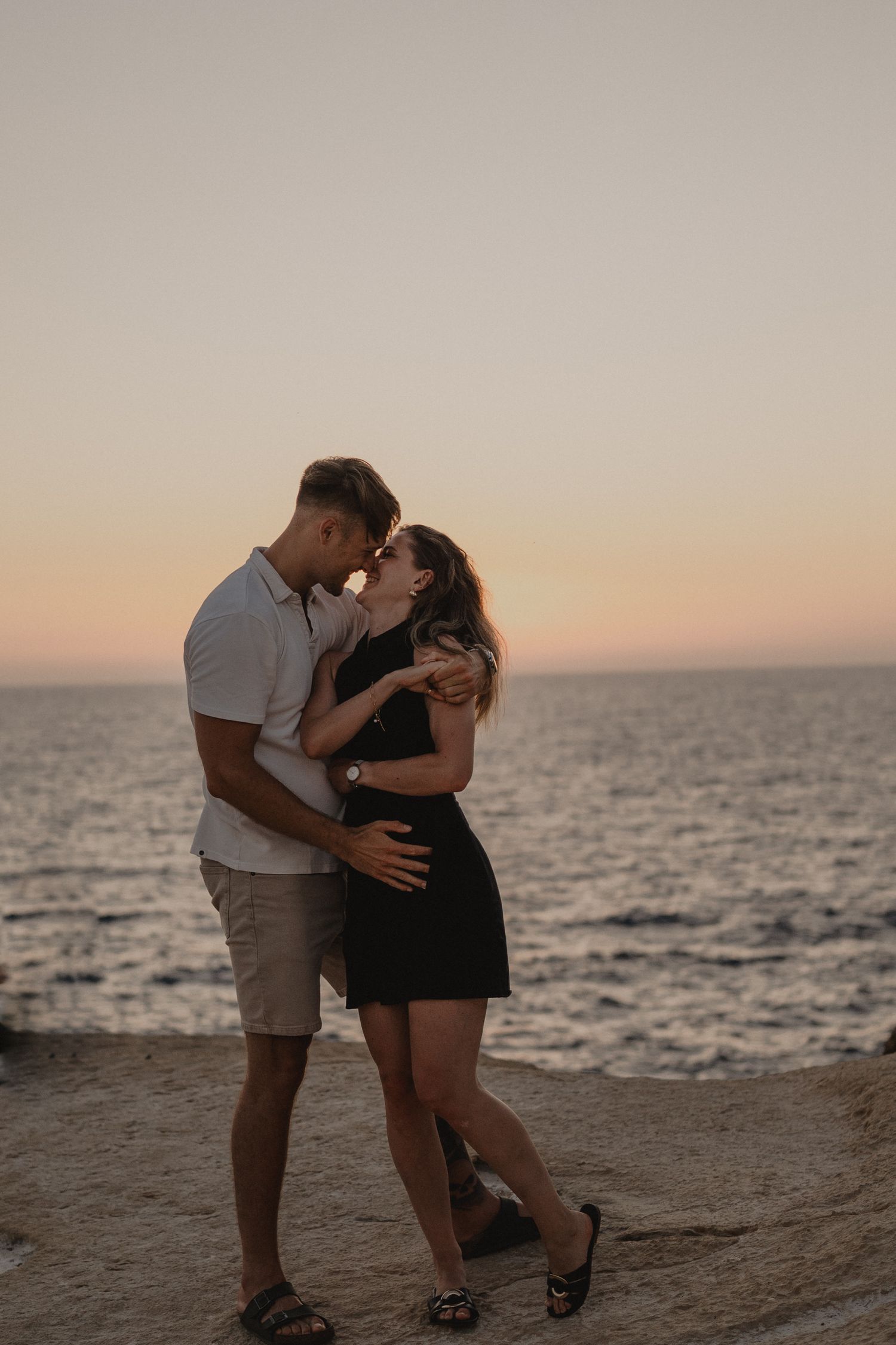 Couple sharing a romantic kiss at sunset on rocky coastal cliffs with ocean waves in the background.