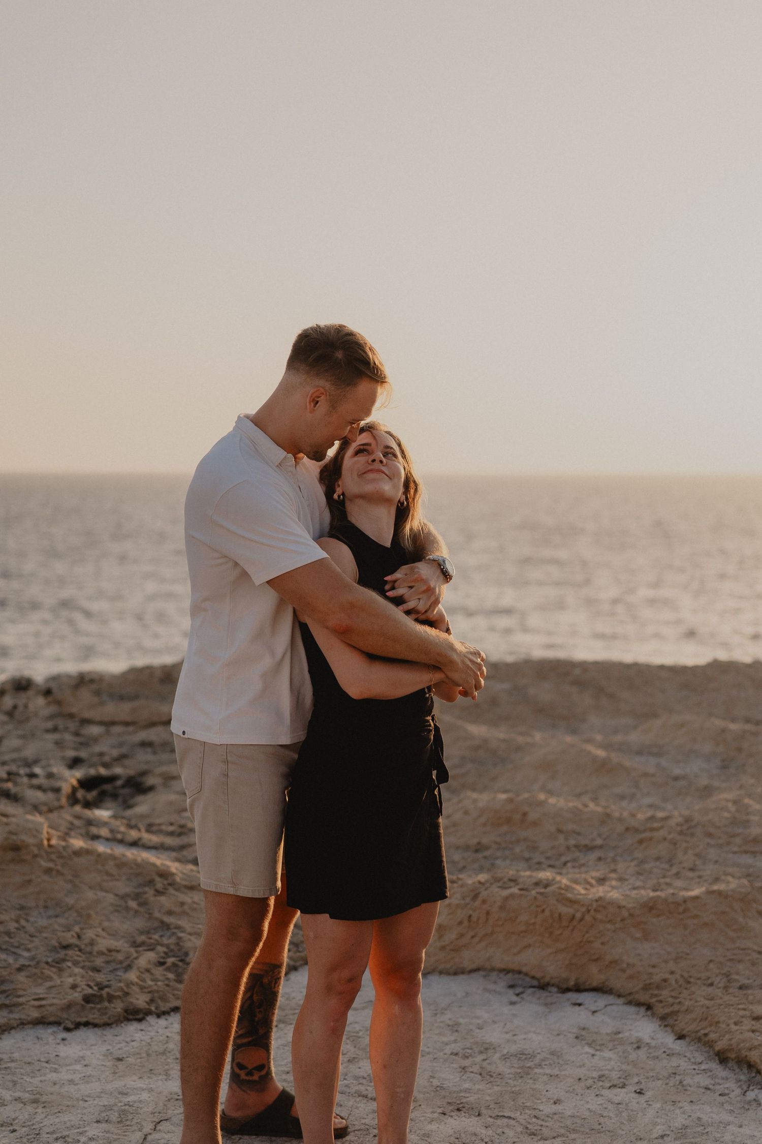 Couple embracing romantically on rocky coastal cliffs at golden sunset, woman in black dress smiling joyfully.
