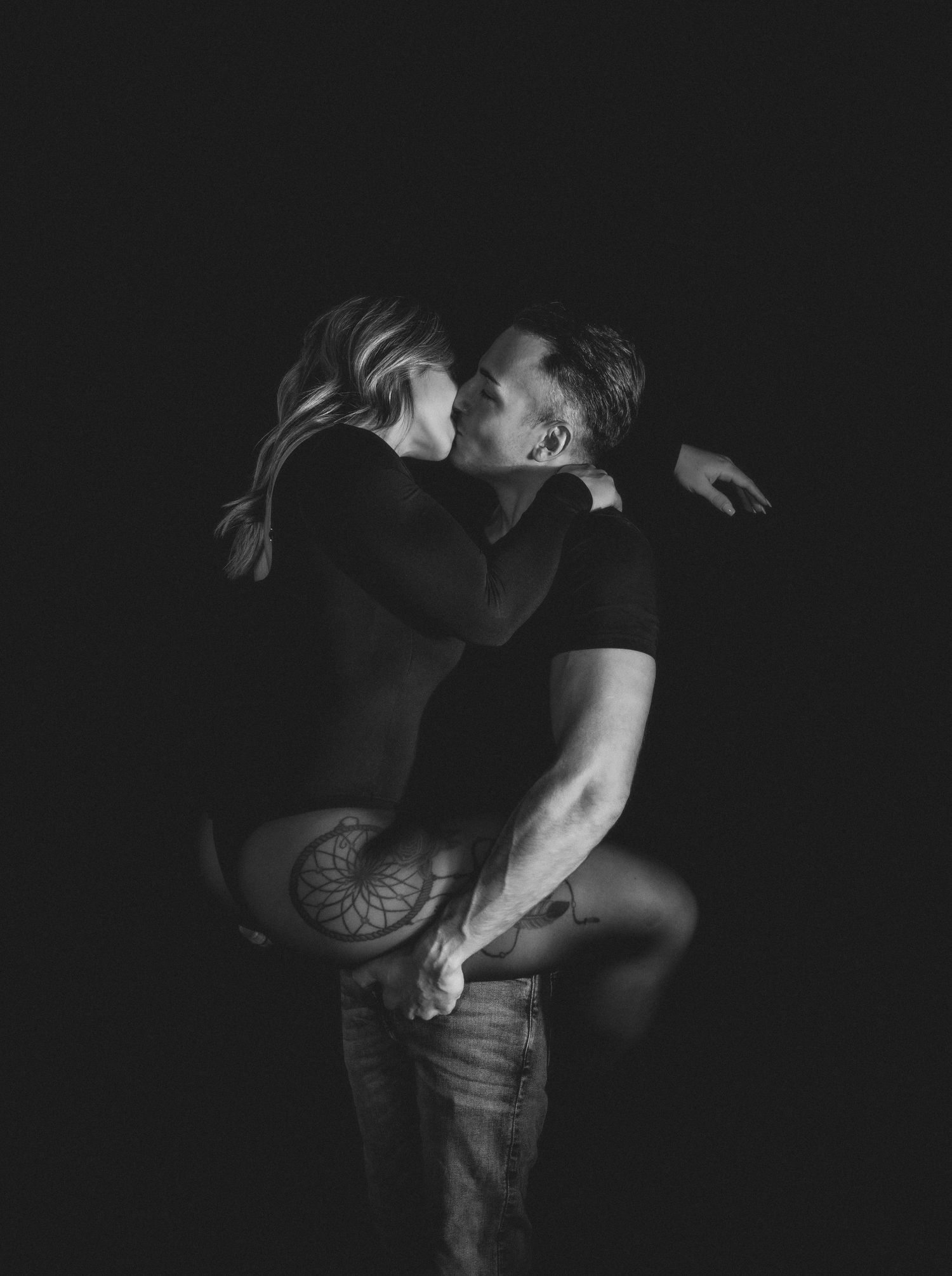 Black and white photo of a couple embracing and kissing passionately against a dark background.