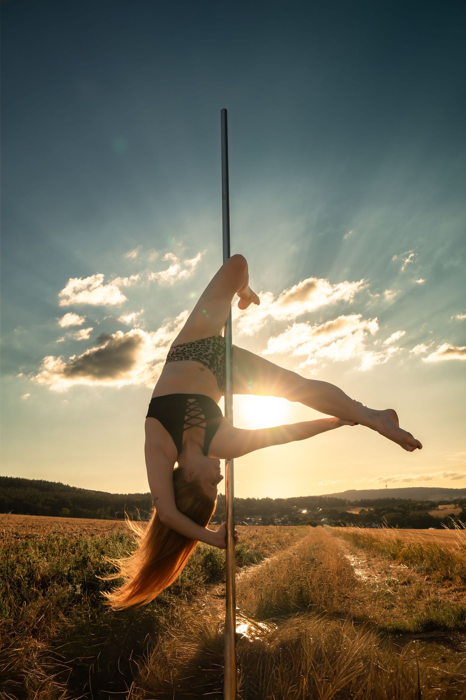 A flexible woman performs an inverted pole dance move outdoors in a golden field at sunset.