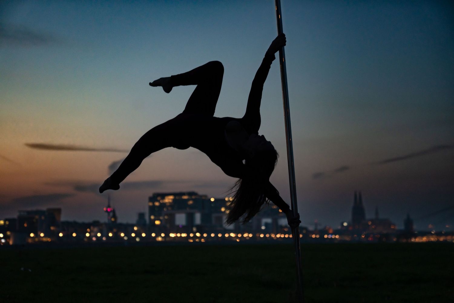 Silhouette of a pole dancer performing an acrobatic move against a twilight city skyline.