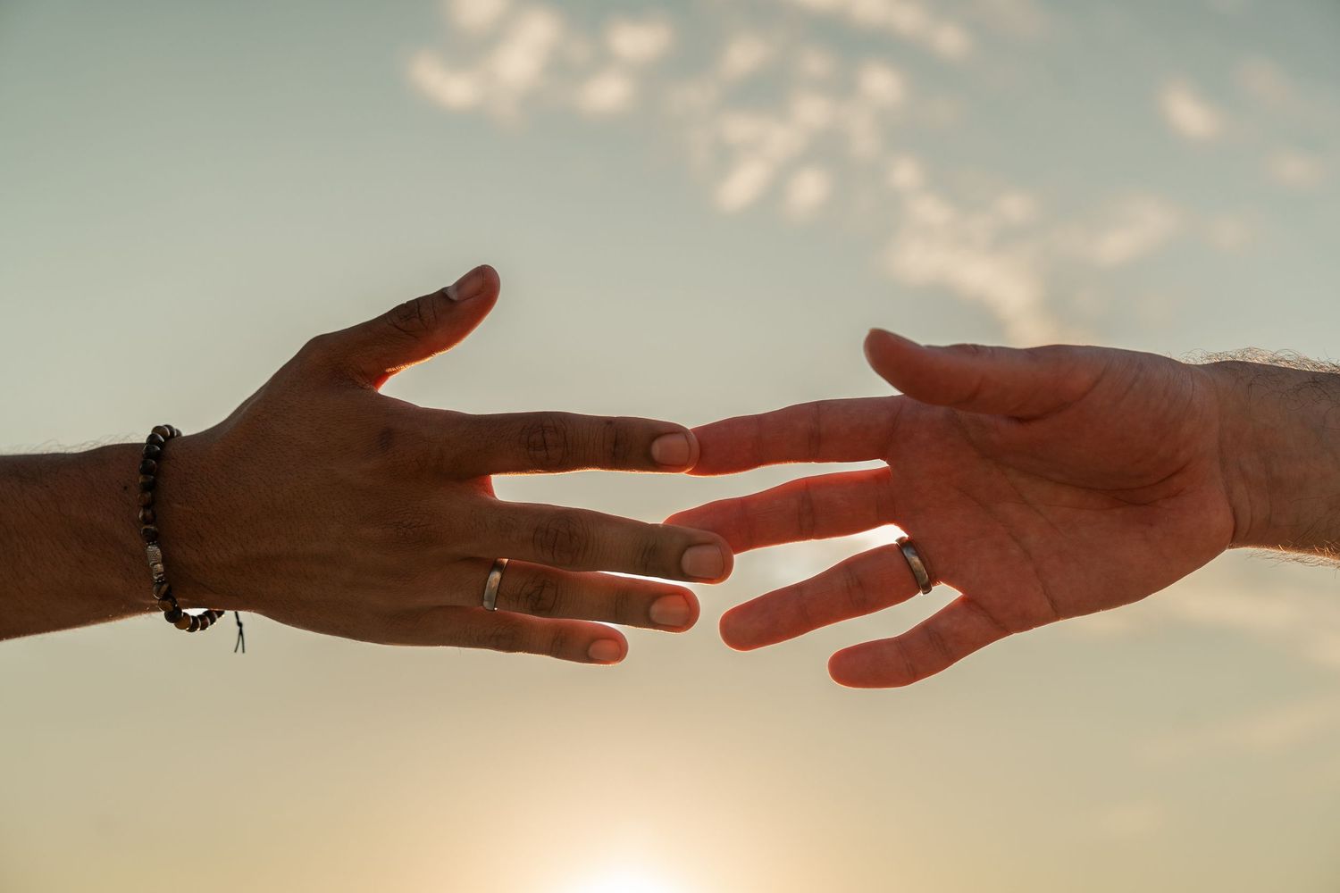 Two hands reaching toward each other against a sunset sky, one wearing a bracelet and ring, symbolizing connection.