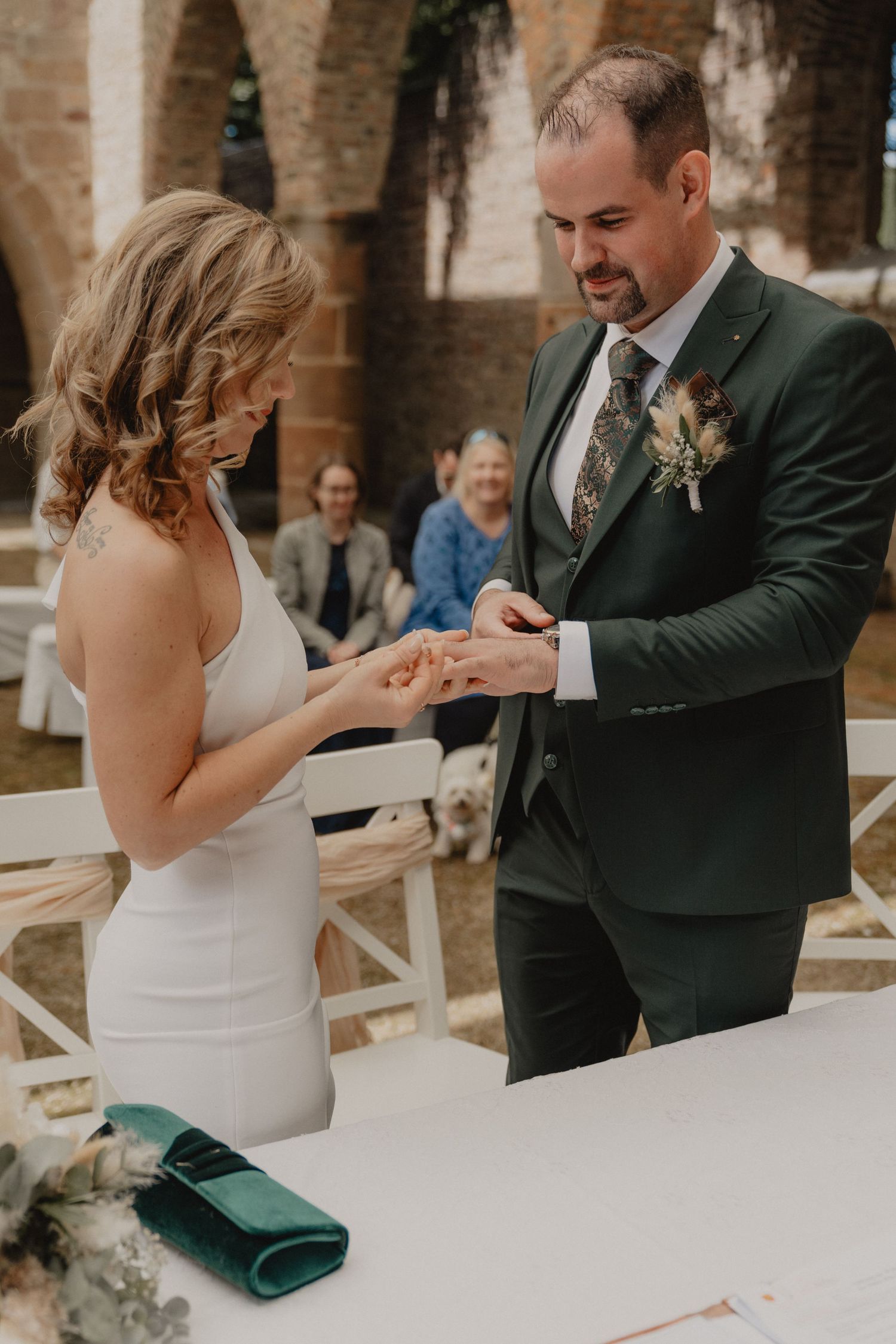 A wedding ring exchange ceremony in front of stone arches with guests watching in the background.