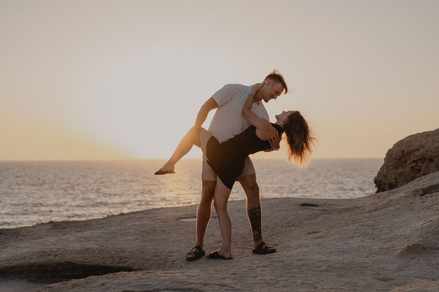 Couple sharing a romantic dip kiss on rocky shore at golden sunset with ocean backdrop.