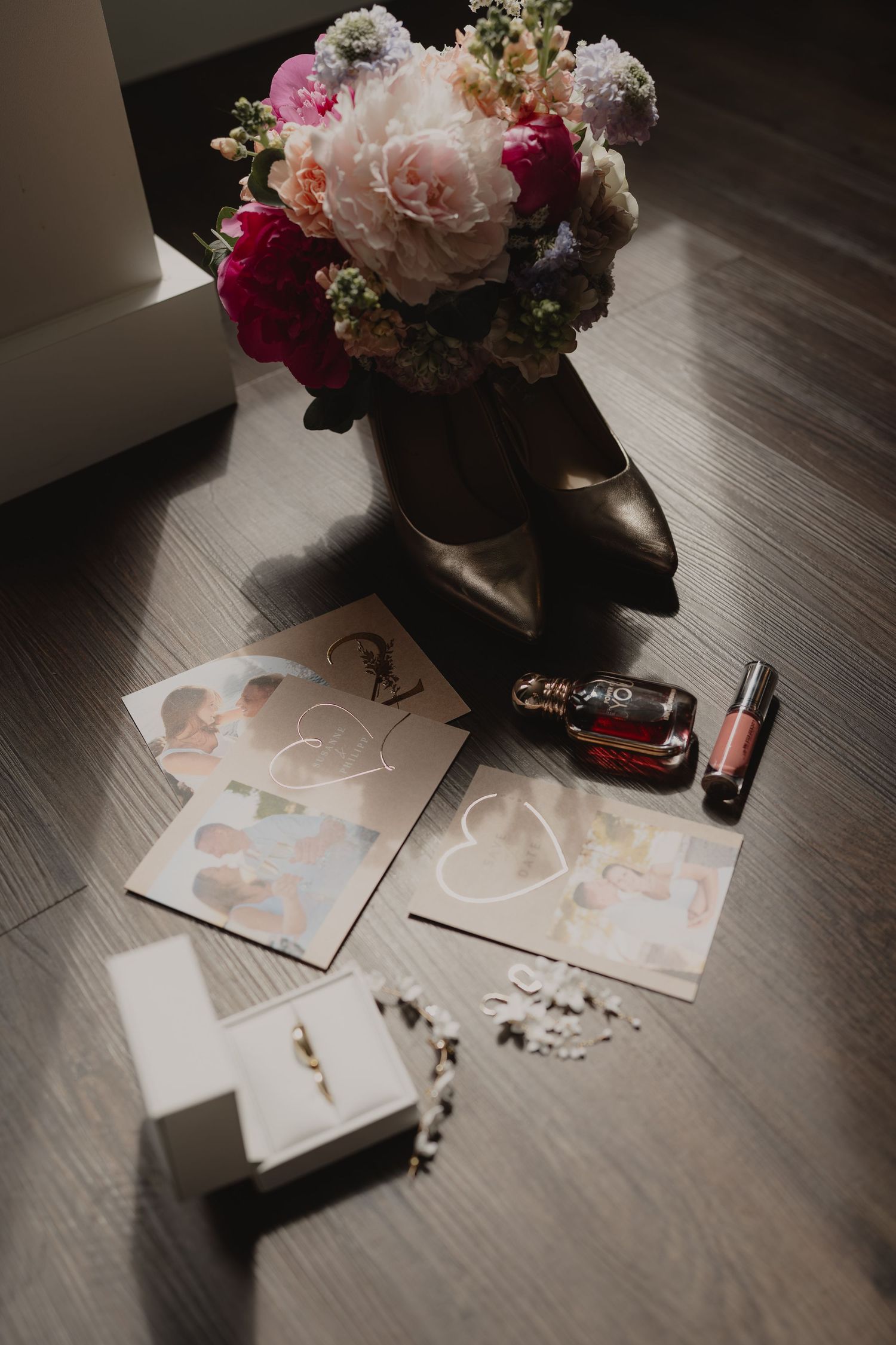 Bridal flat lay with floral bouquet, heeled shoes, wedding cards, perfume, lipstick, jewelry box, and rings on wood floor.