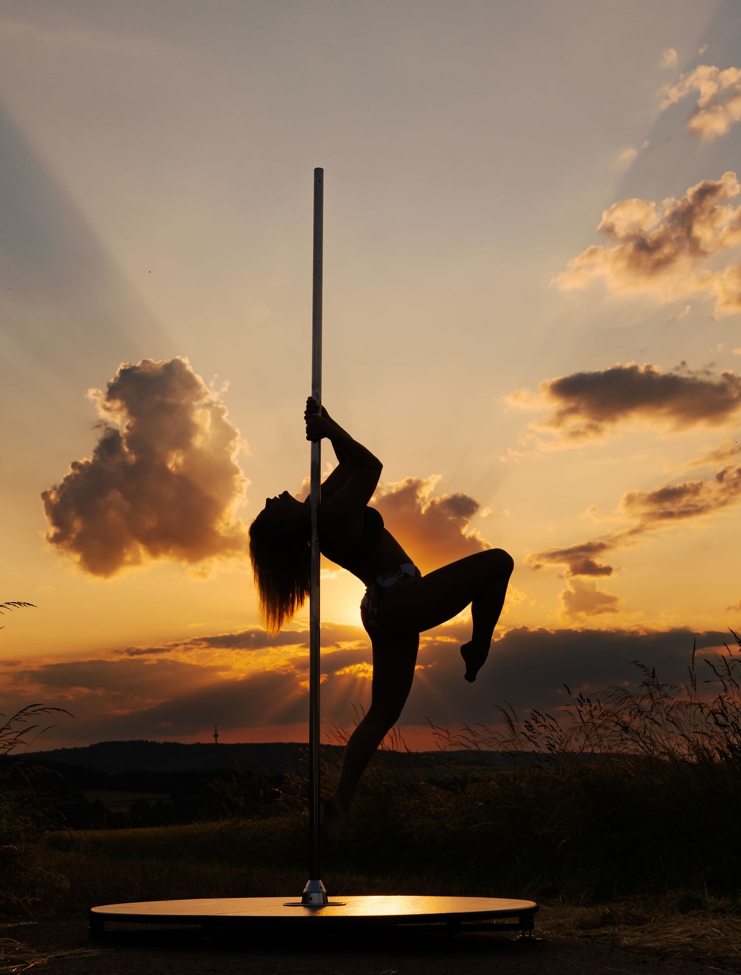 Silhouette of a woman pole dancing outdoors against a dramatic golden sunset sky with clouds.