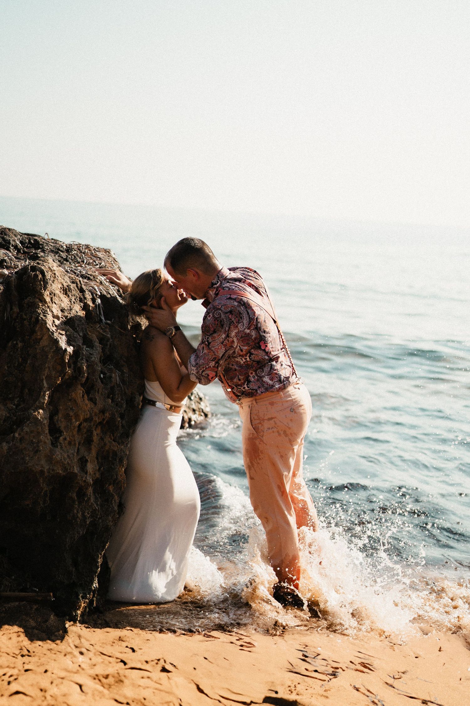 Couple shares intimate moment by rocky beach as waves splash dramatically behind them.