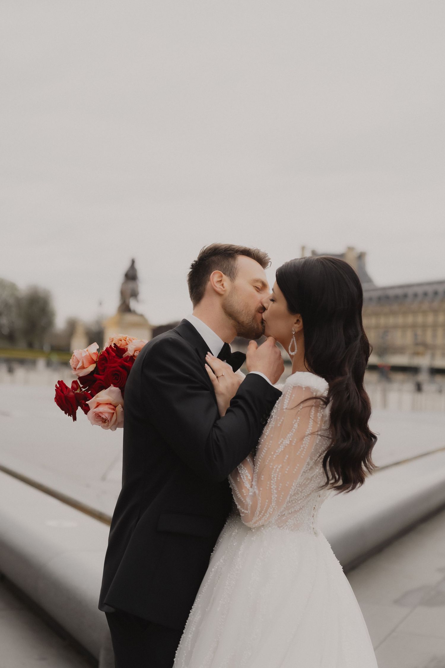 Romantic wedding couple sharing a kiss against a cloudy sky with historic architecture in the background.