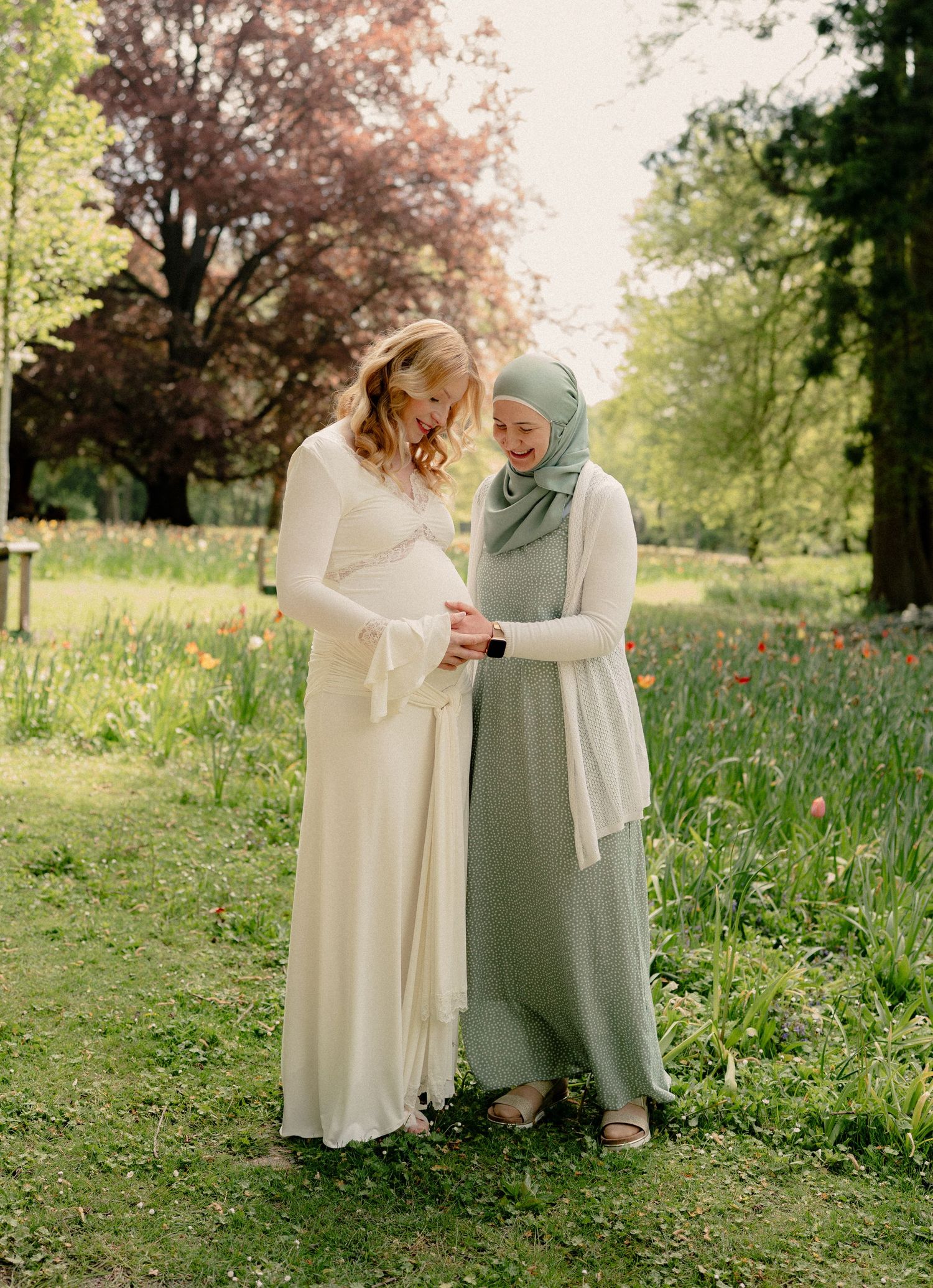 Two women in elegant dresses stand together in a garden, one pregnant in white, the other in sage green hijab.