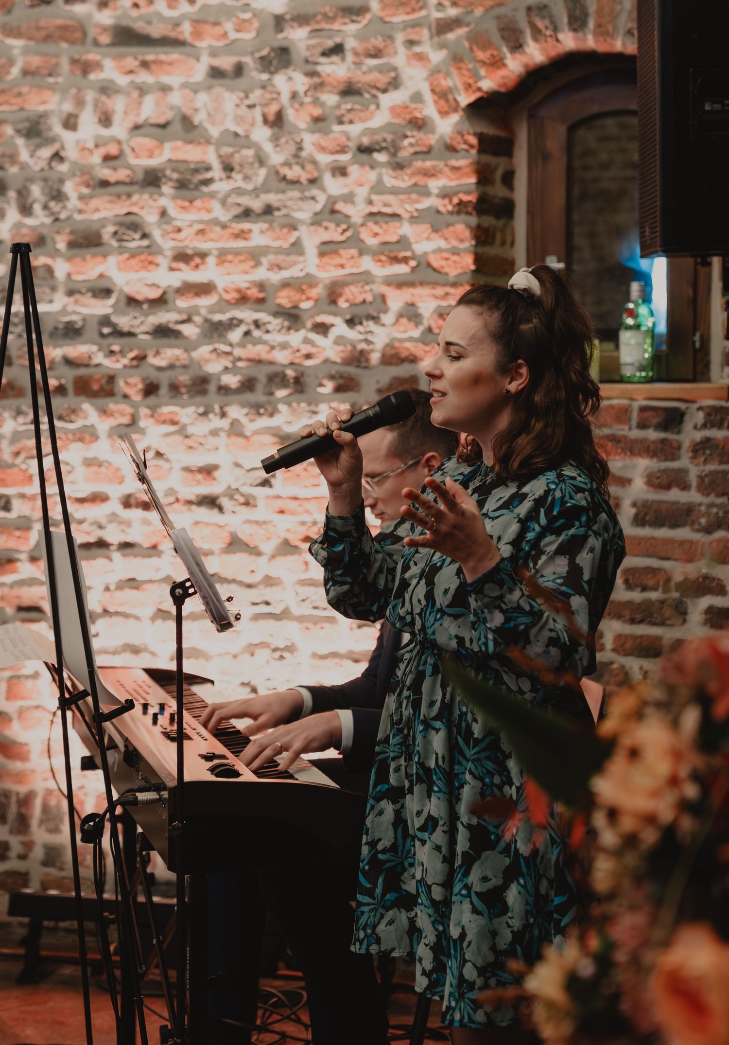 Singer in floral dress performs with keyboard at rustic brick venue.