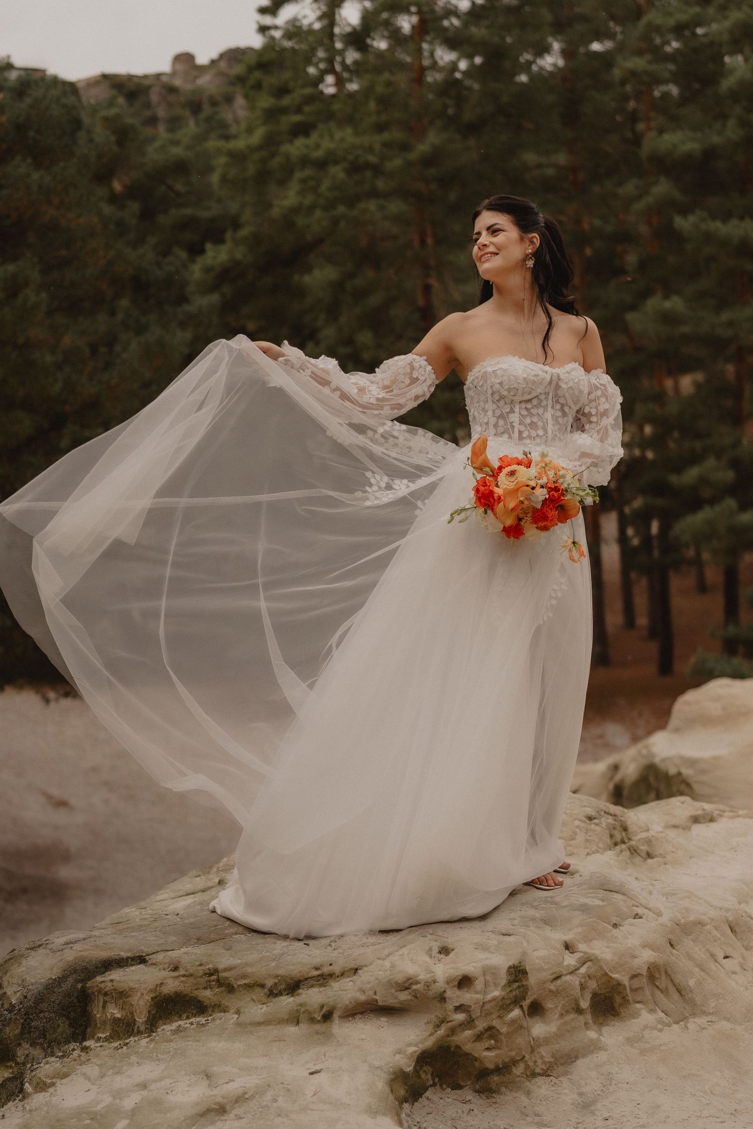 A bride in a flowing white off-shoulder gown holds an orange bouquet while standing on rocky terrain in a forest setting.