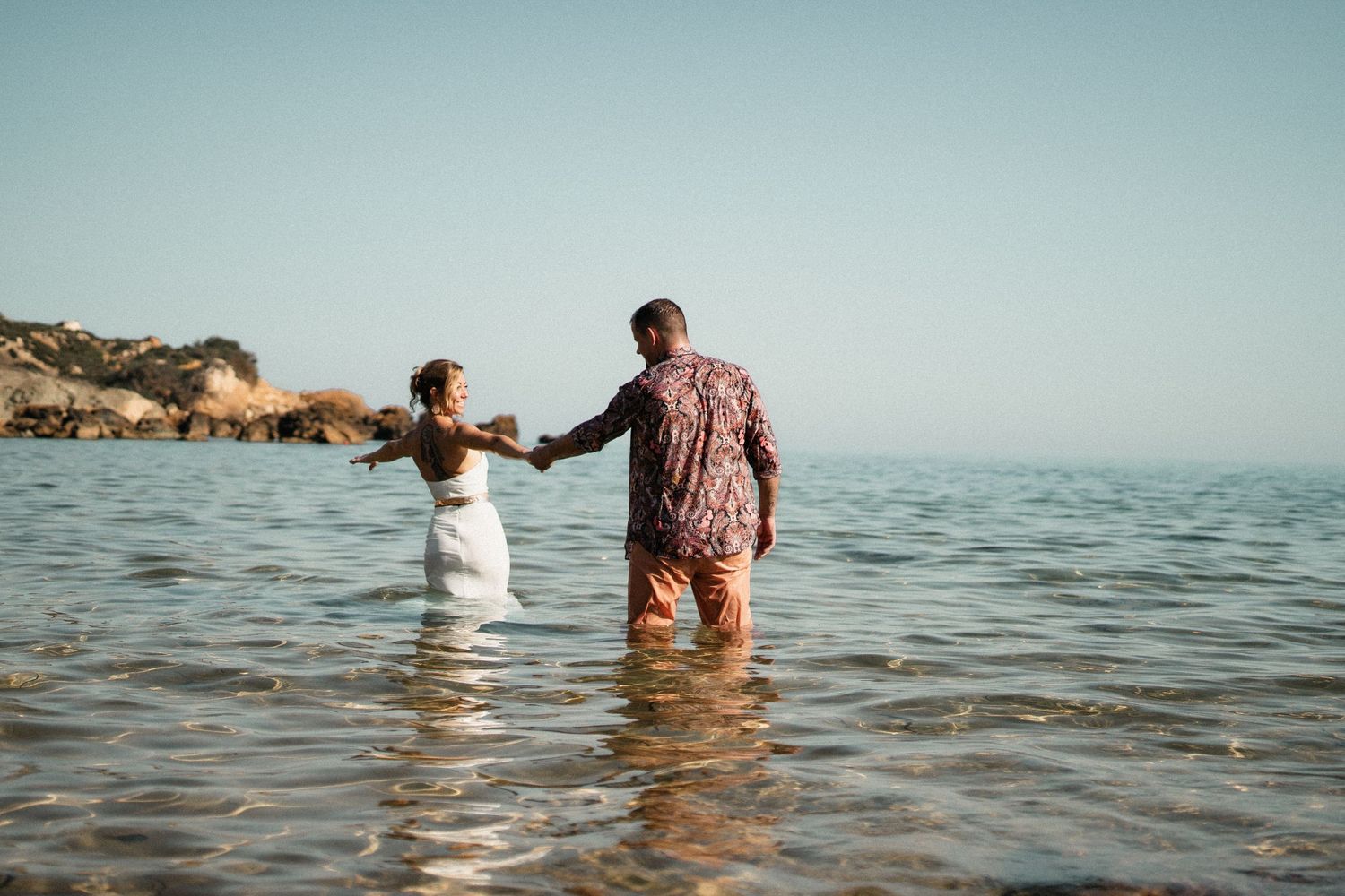 Couple holding hands while walking together in shallow ocean water during sunset at the beach.