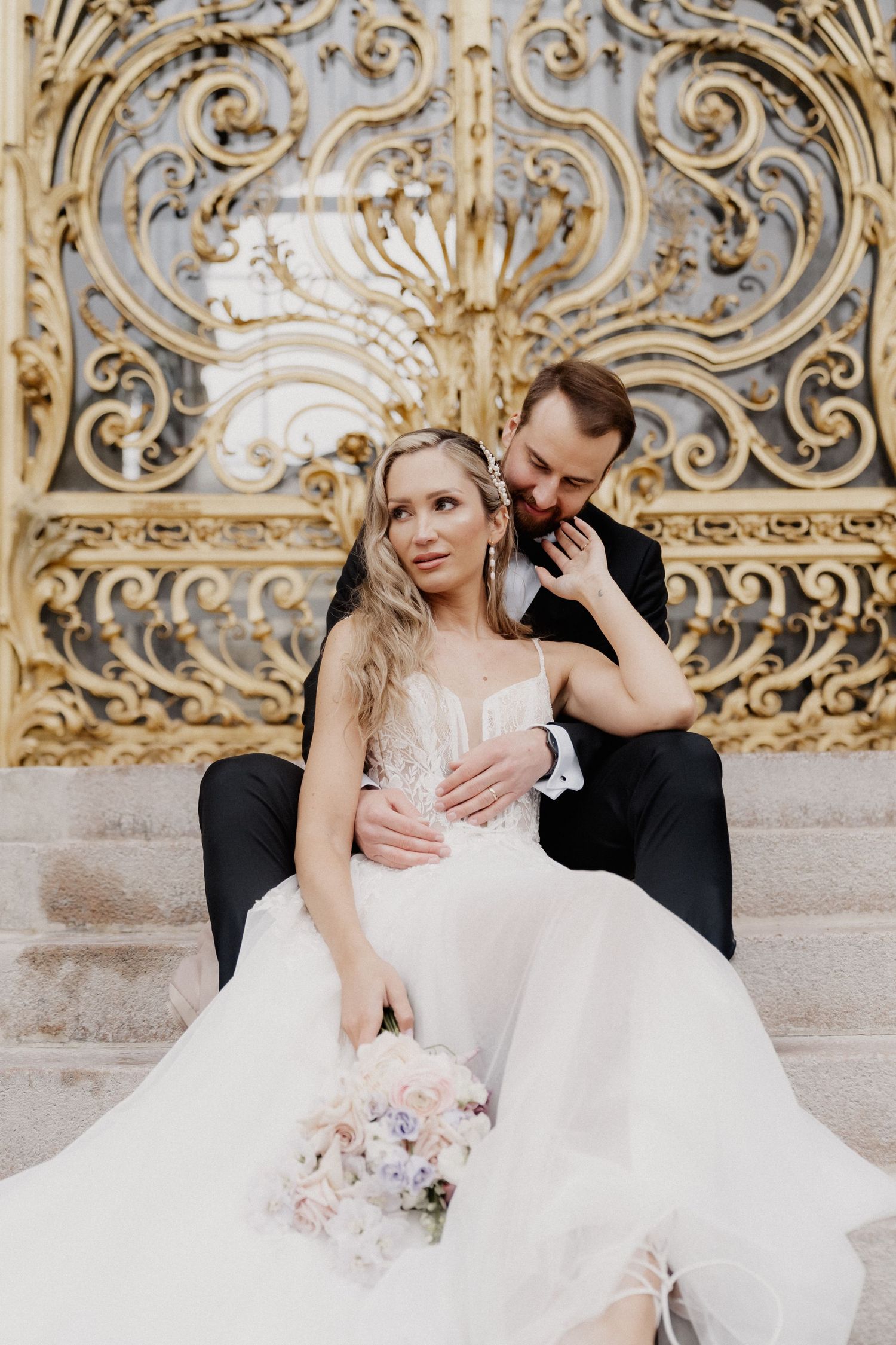 A romantic wedding couple sits together against an ornate baroque-style wall with gold scrollwork details.