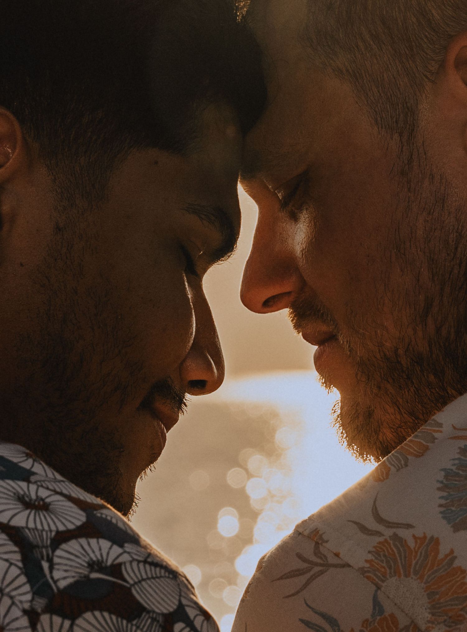 Two men with beards face each other intimately in warm golden backlight, wearing floral shirts.