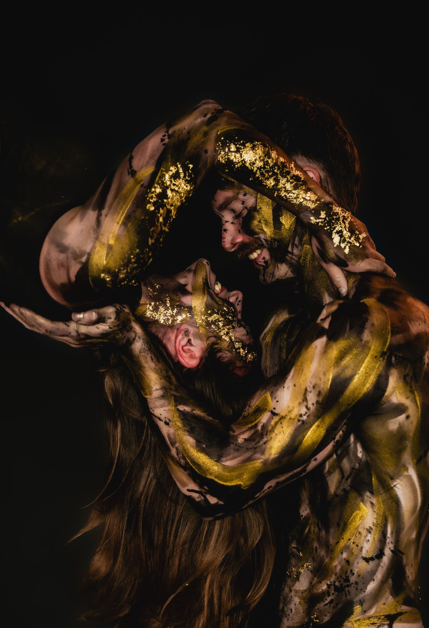 Two figures covered in gold paint and leaf embrace dramatically against a dark background in an artistic performance.