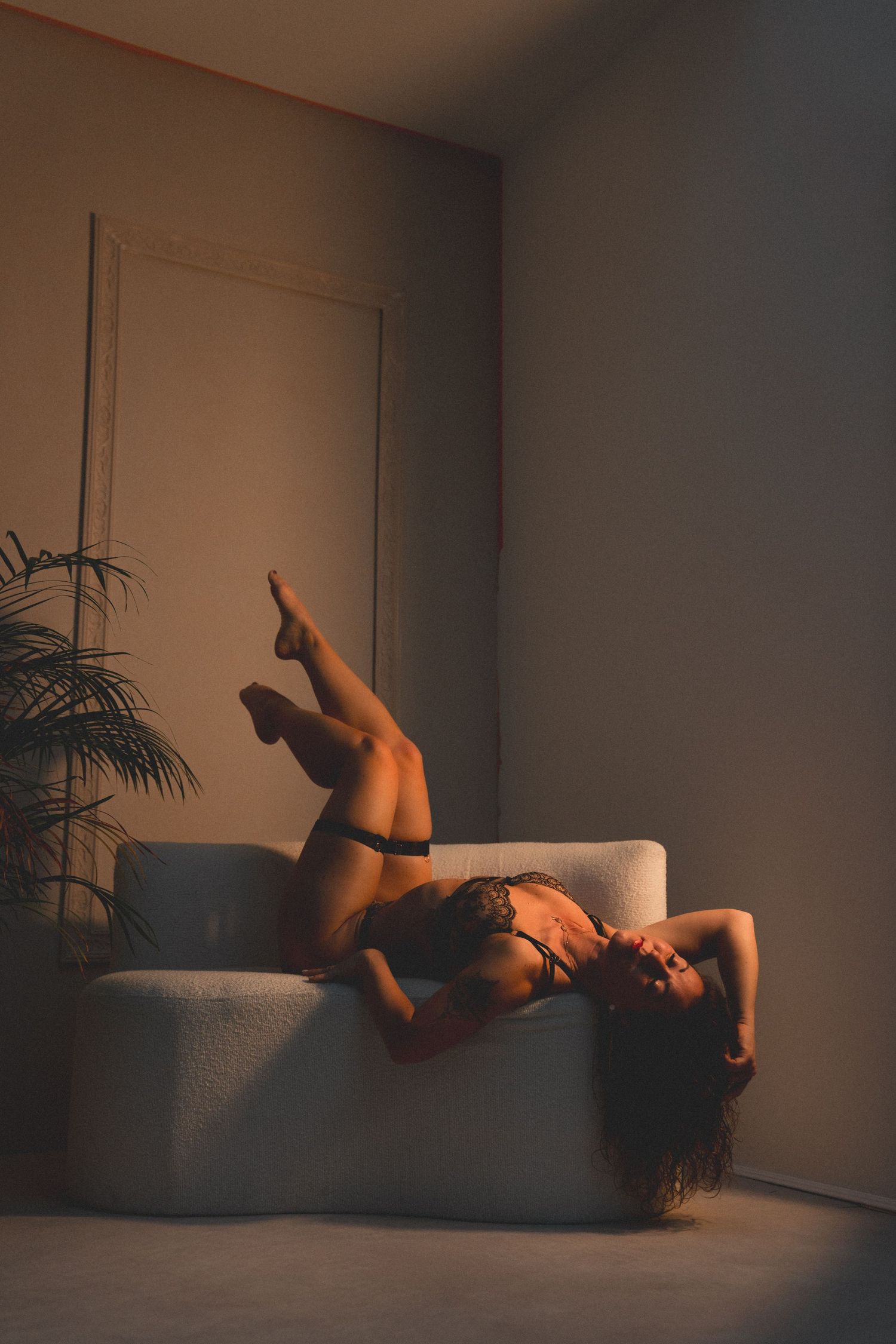 Woman in lingerie posing on a white sofa in warm moody lighting with a plant in the background.