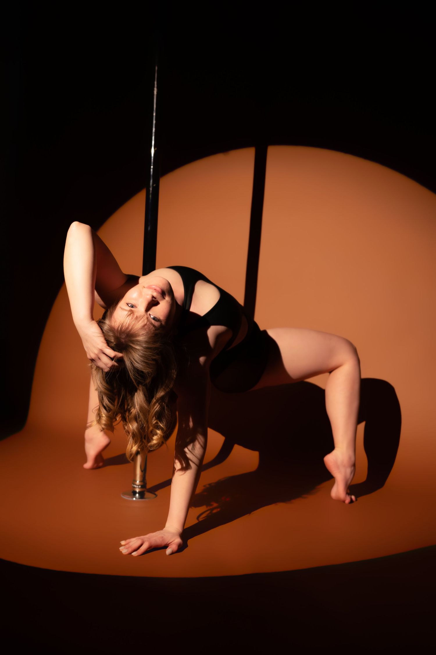 A pole dancer in a black bodysuit performs a dramatic backbend pose under a spotlight on a brown studio background.
