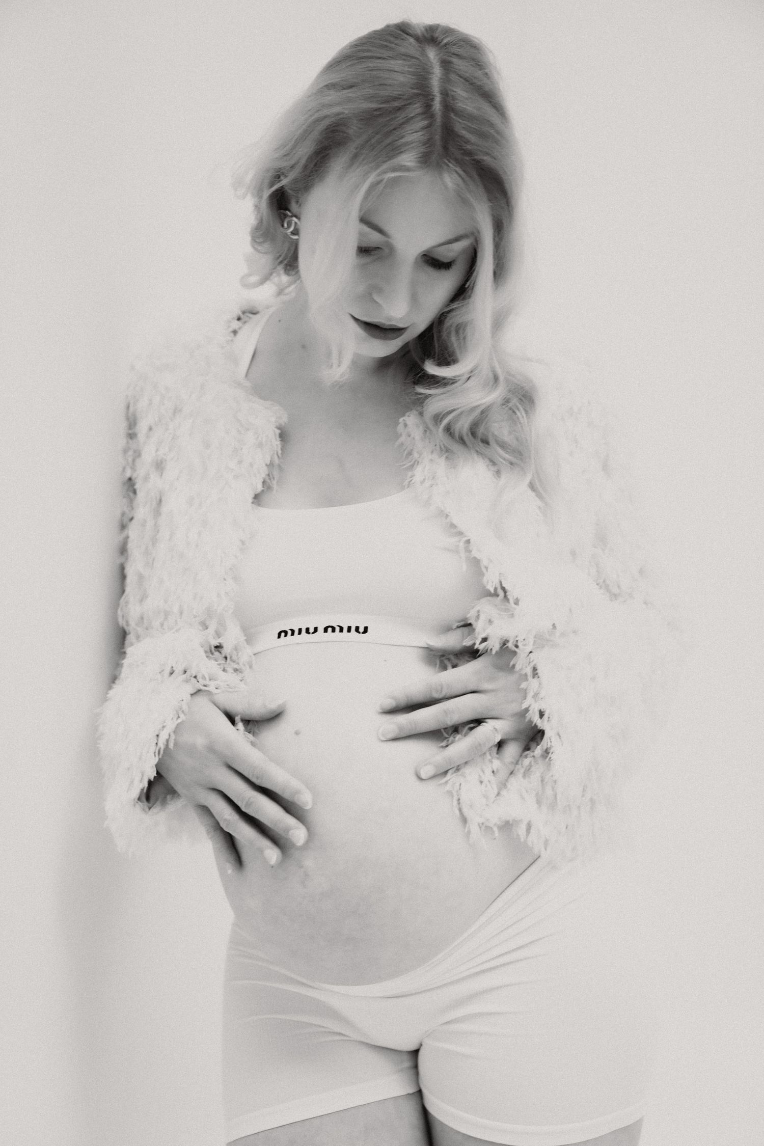 Black and white photo of a pregnant blonde woman in white shorts and fluffy jacket cradling her baby bump.