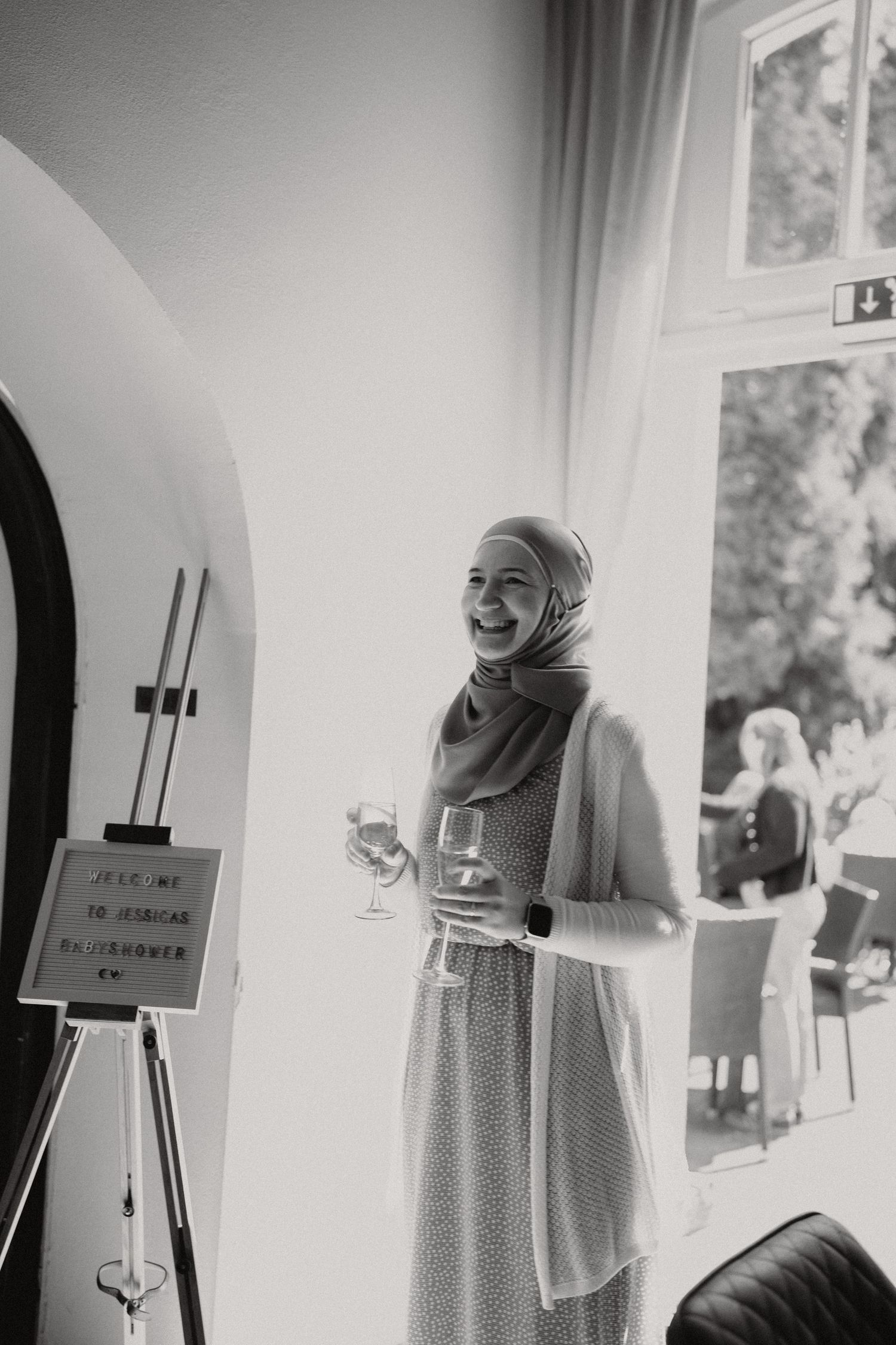 Black and white photo of a smiling woman in hijab holding a wine glass at an event.