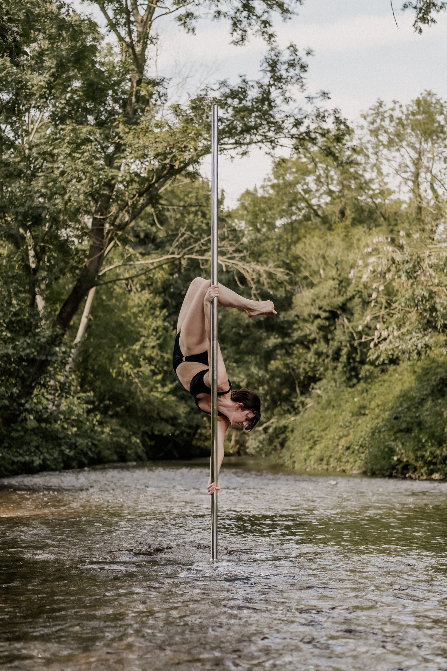 A woman performs an inverted pole dance move outdoors on a metal pole set in a shallow river surrounded by lush green trees.