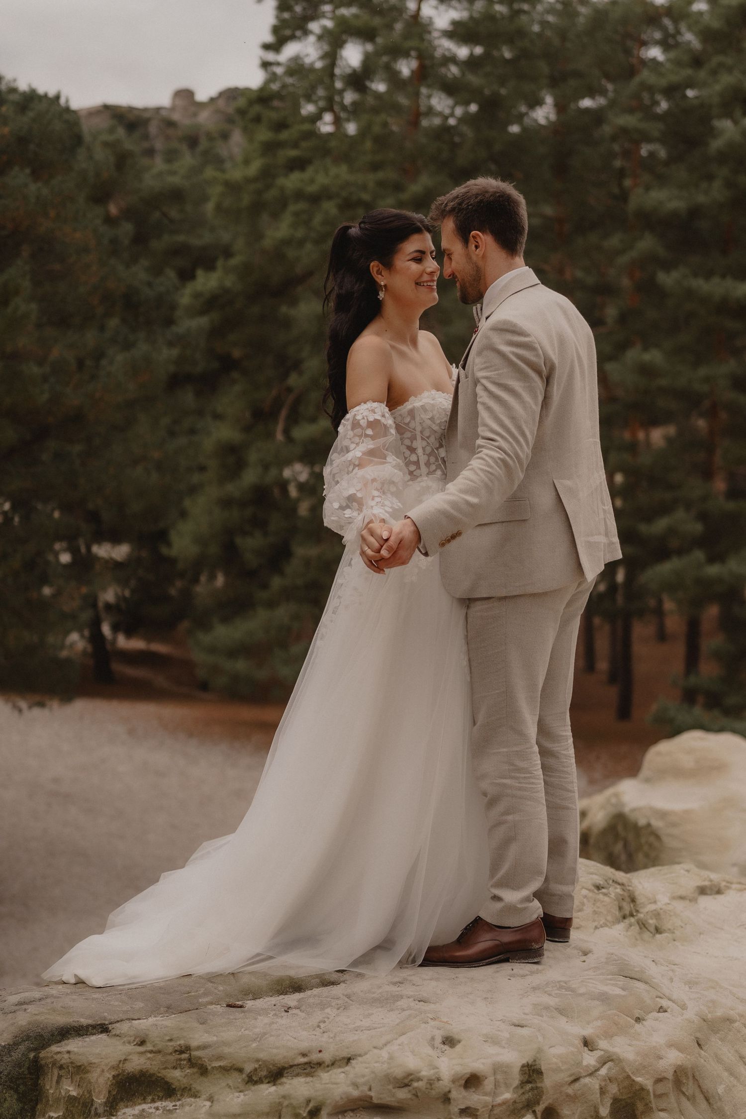 Wedding couple poses together on rocky terrain surrounded by pine trees.