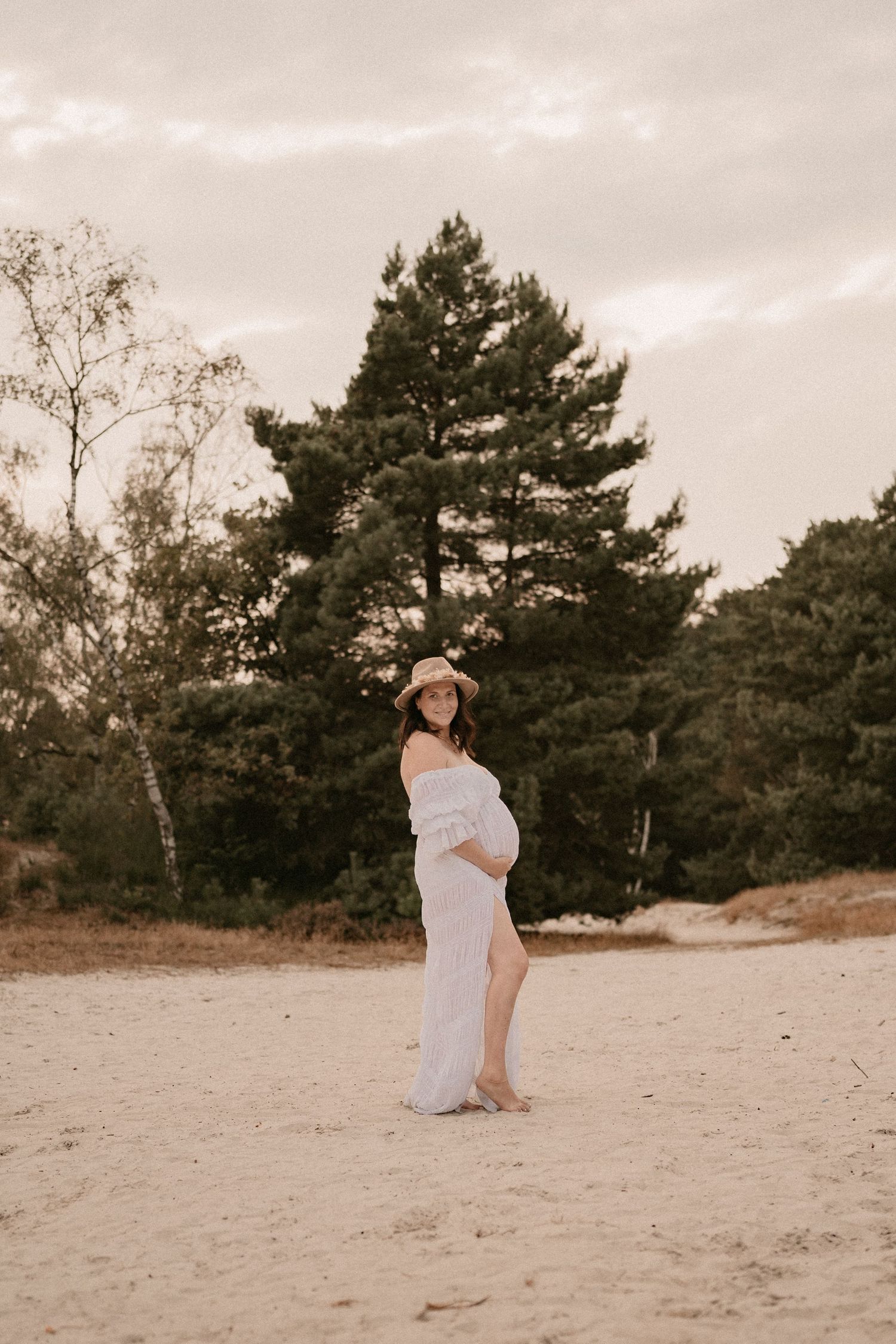 Woman in a flowing white dress and sun hat poses gracefully on a sandy beach.