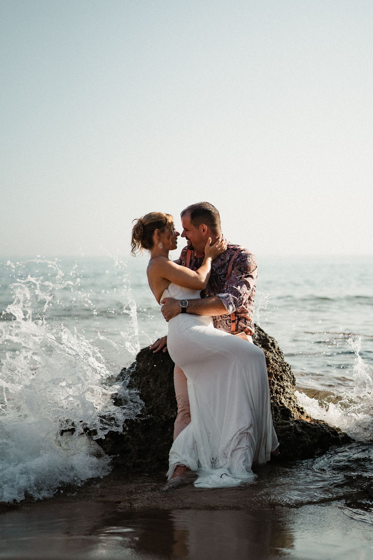 Ocean waves crash around couple seated on rocky outcrop during romantic coastal photoshoot.