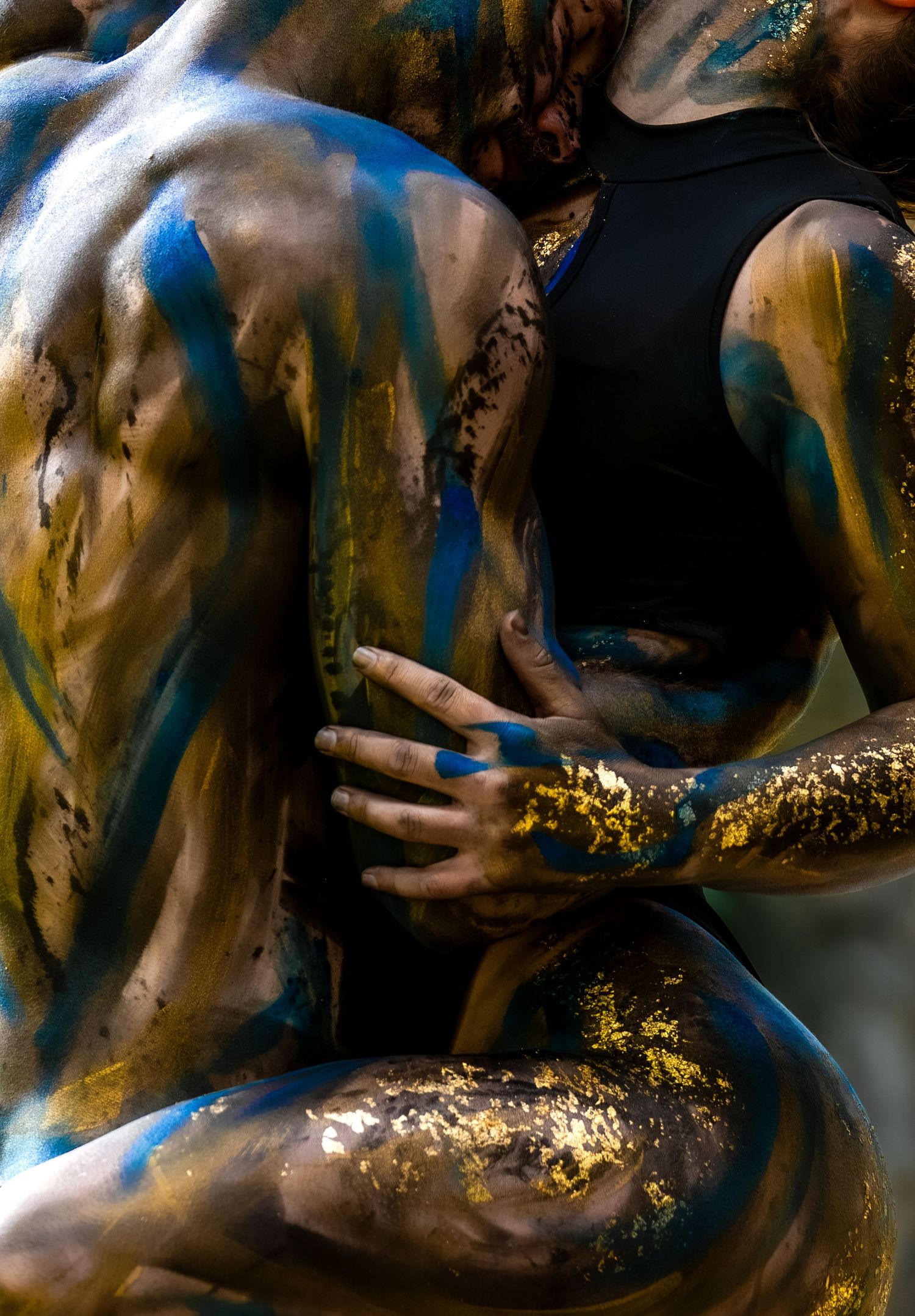 Two figures covered in blue, gold, and brown body paint embrace in an artistic and expressive pose.