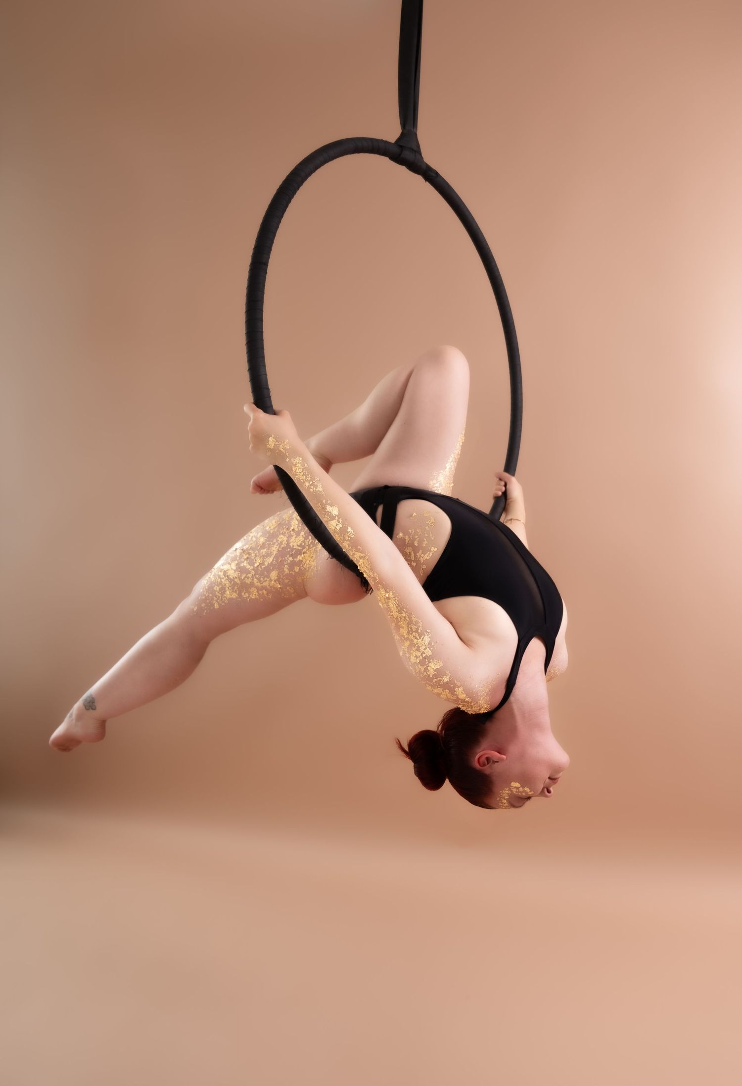 Aerial hoop performer in black leotard executing a graceful backbend pose on a lyra ring against beige background.
