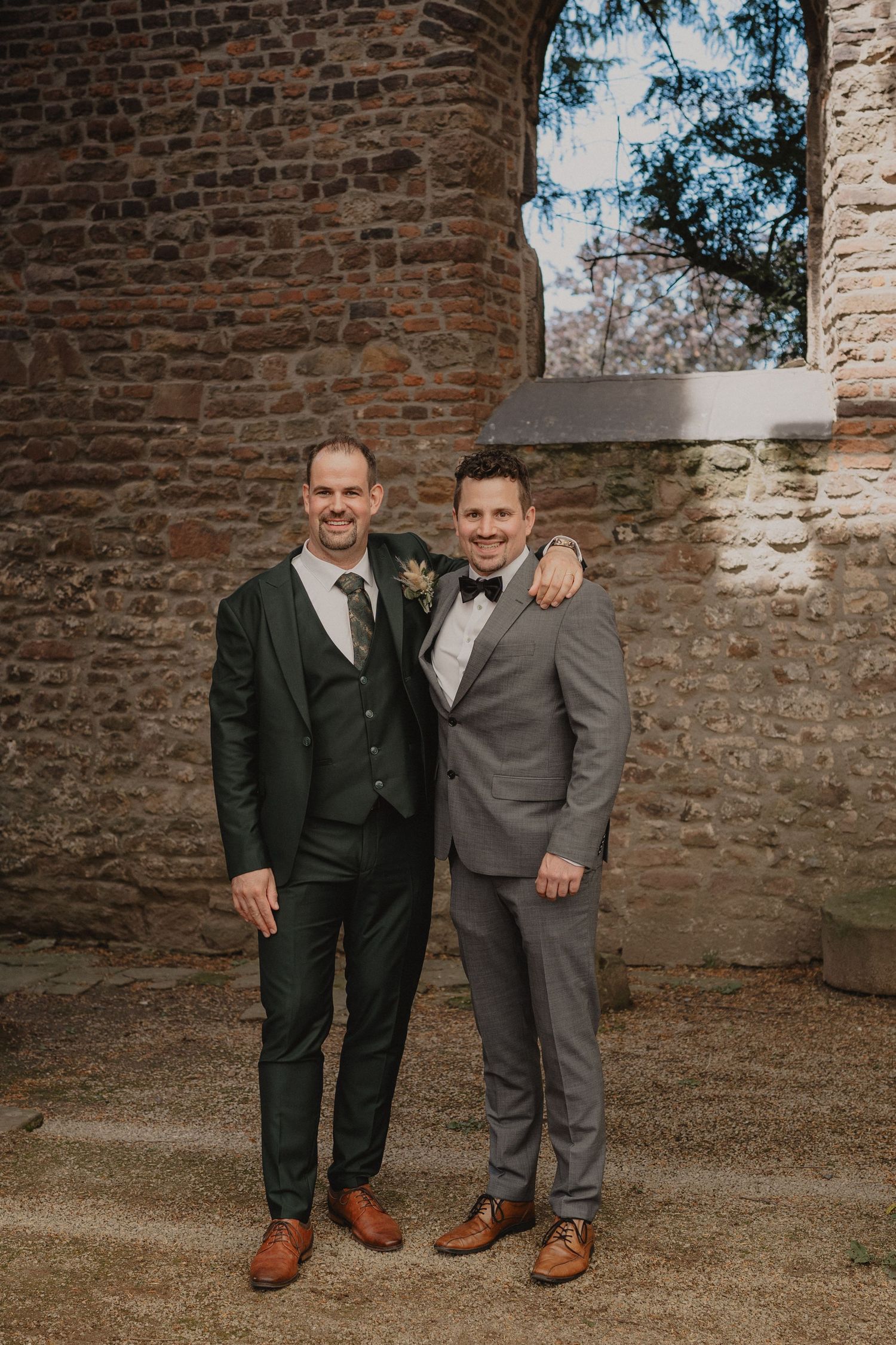 Two men in formal suits posing together against stone wall background at wedding venue.
