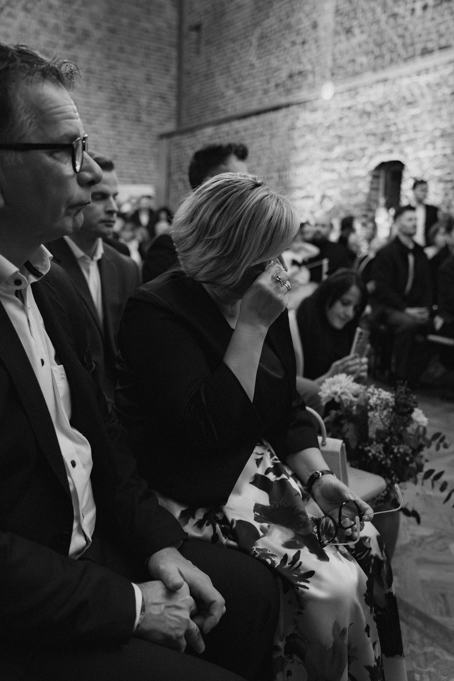 Wedding guests seated in a rustic brick venue with flowers and formal attire.