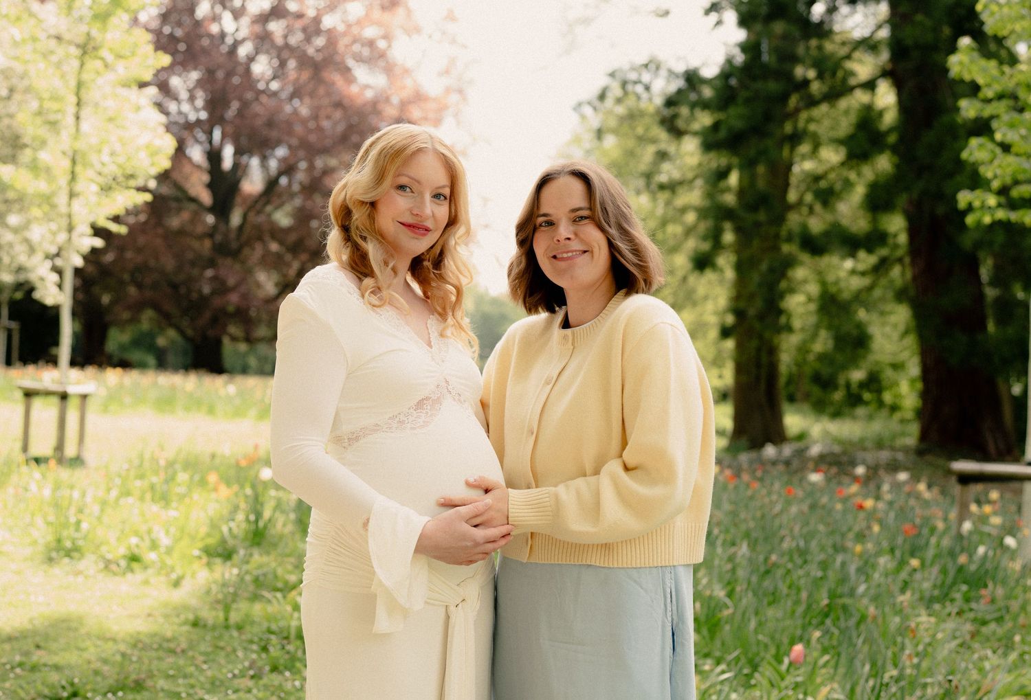 Two women smiling outdoors in a garden, one pregnant in white dress, other in yellow cardigan and blue skirt.