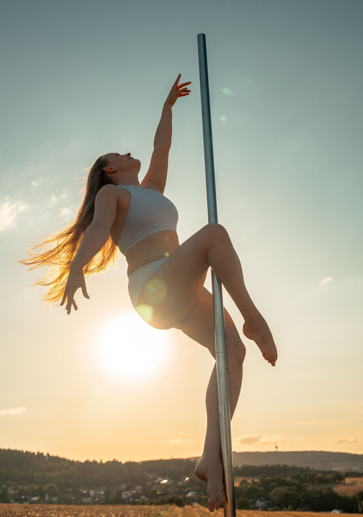 Woman performing pole dance outdoors at sunset, gracefully extending her arm upward against a golden sky.