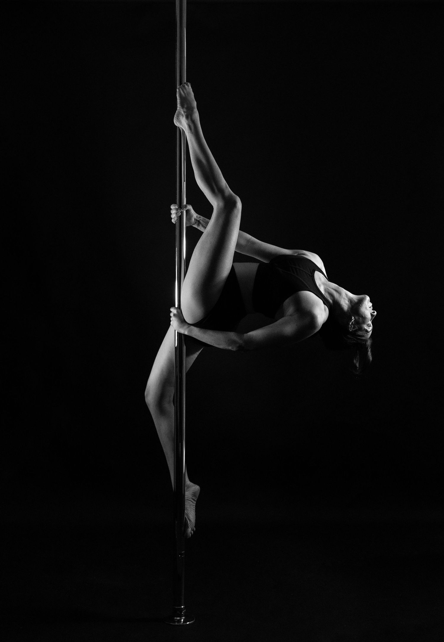 Black and white photo of a woman performing an athletic pole dance pose, arching gracefully against a dark background.