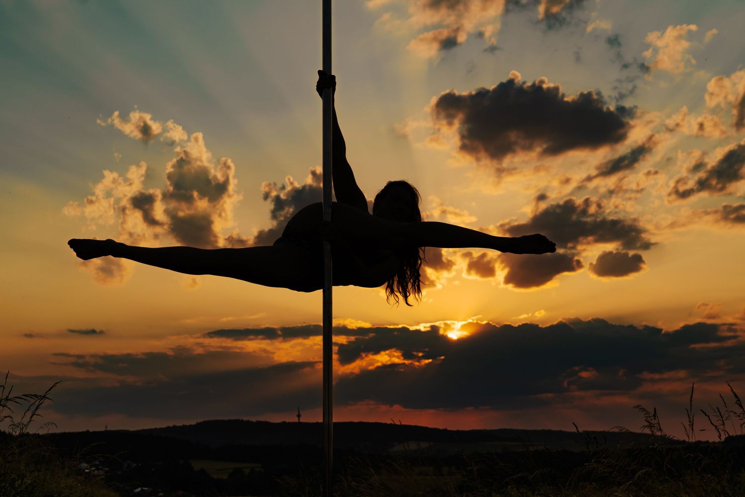 Silhouette of a woman performing a split on a pole against a dramatic golden sunset sky with clouds.