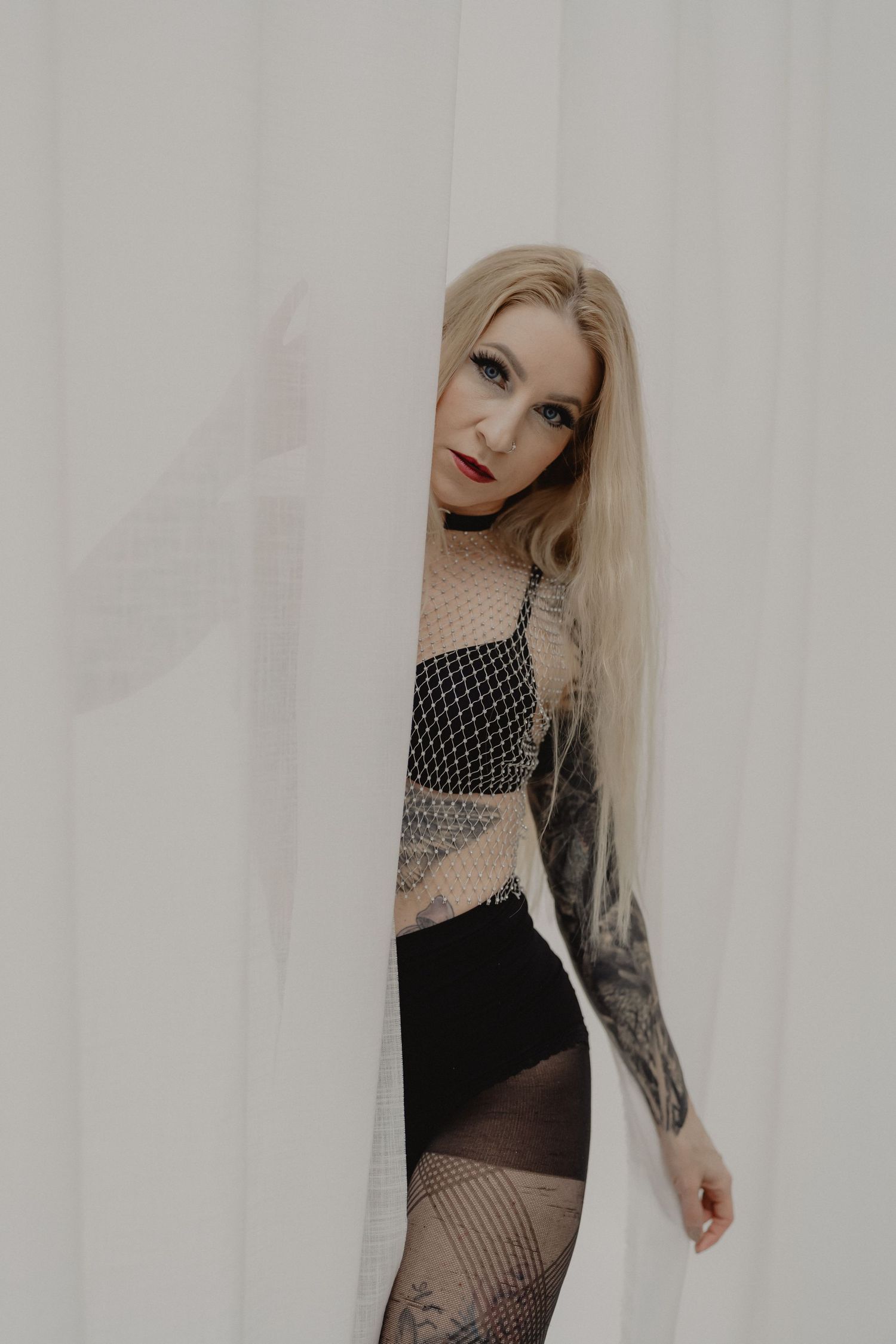 Gothic model in black lingerie and fishnet stockings poses against white wall showing tattoo sleeve.