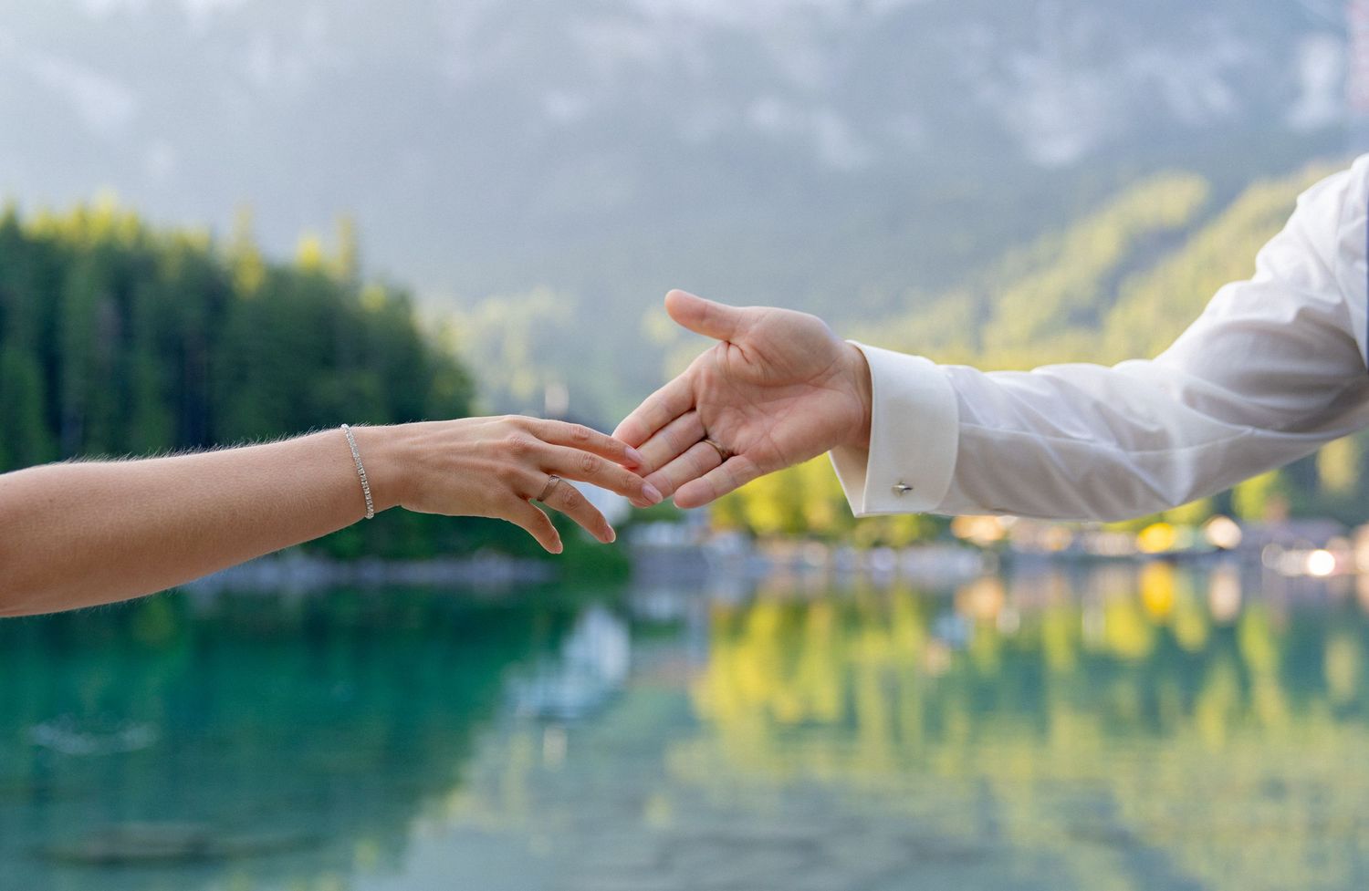 Hand reaches out to touch the calm surface of a pristine mountain lake surrounded by evergreen forest and peaks.