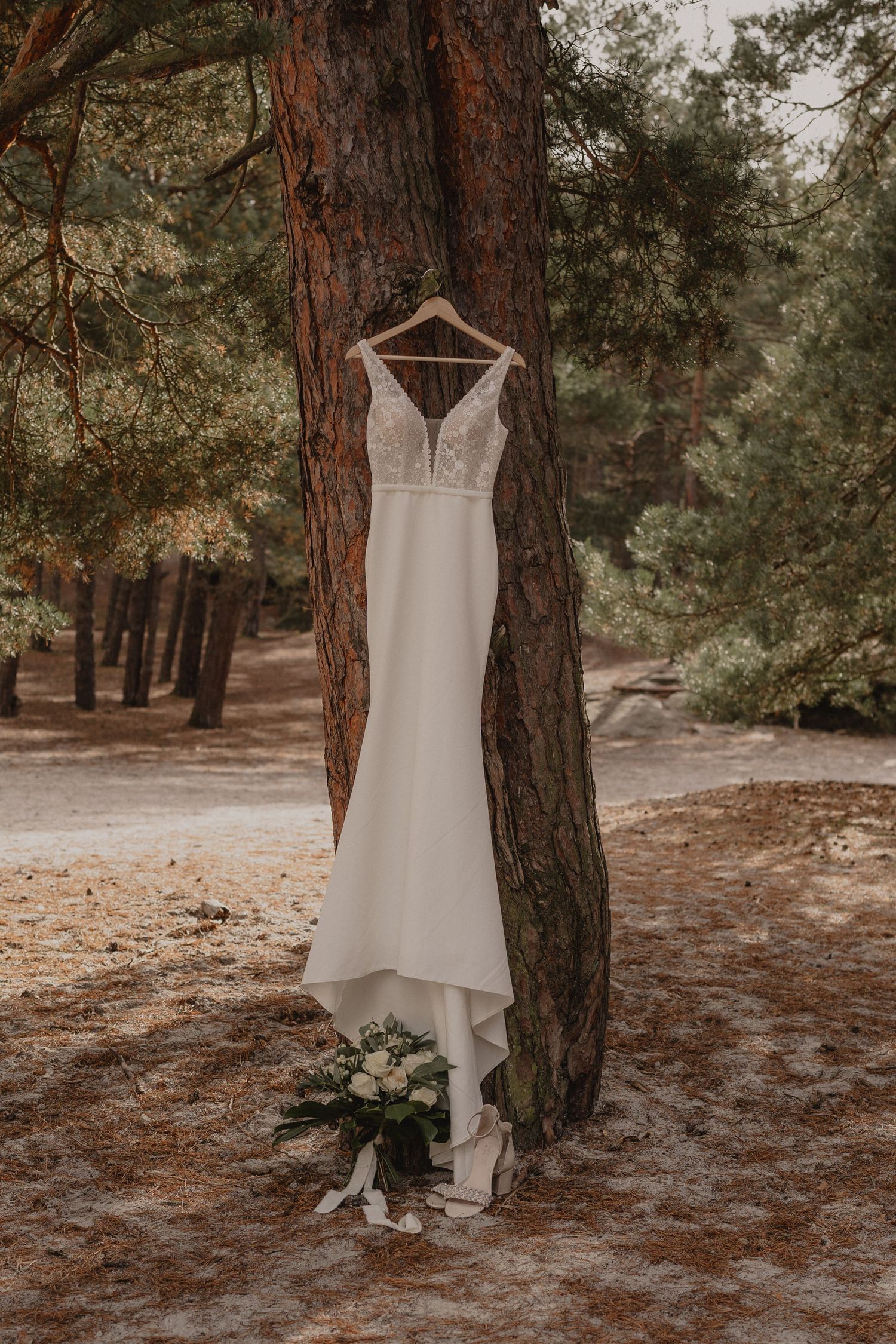 White wedding dress hangs elegantly against pine trees in a forest setting with scattered pine needles below.