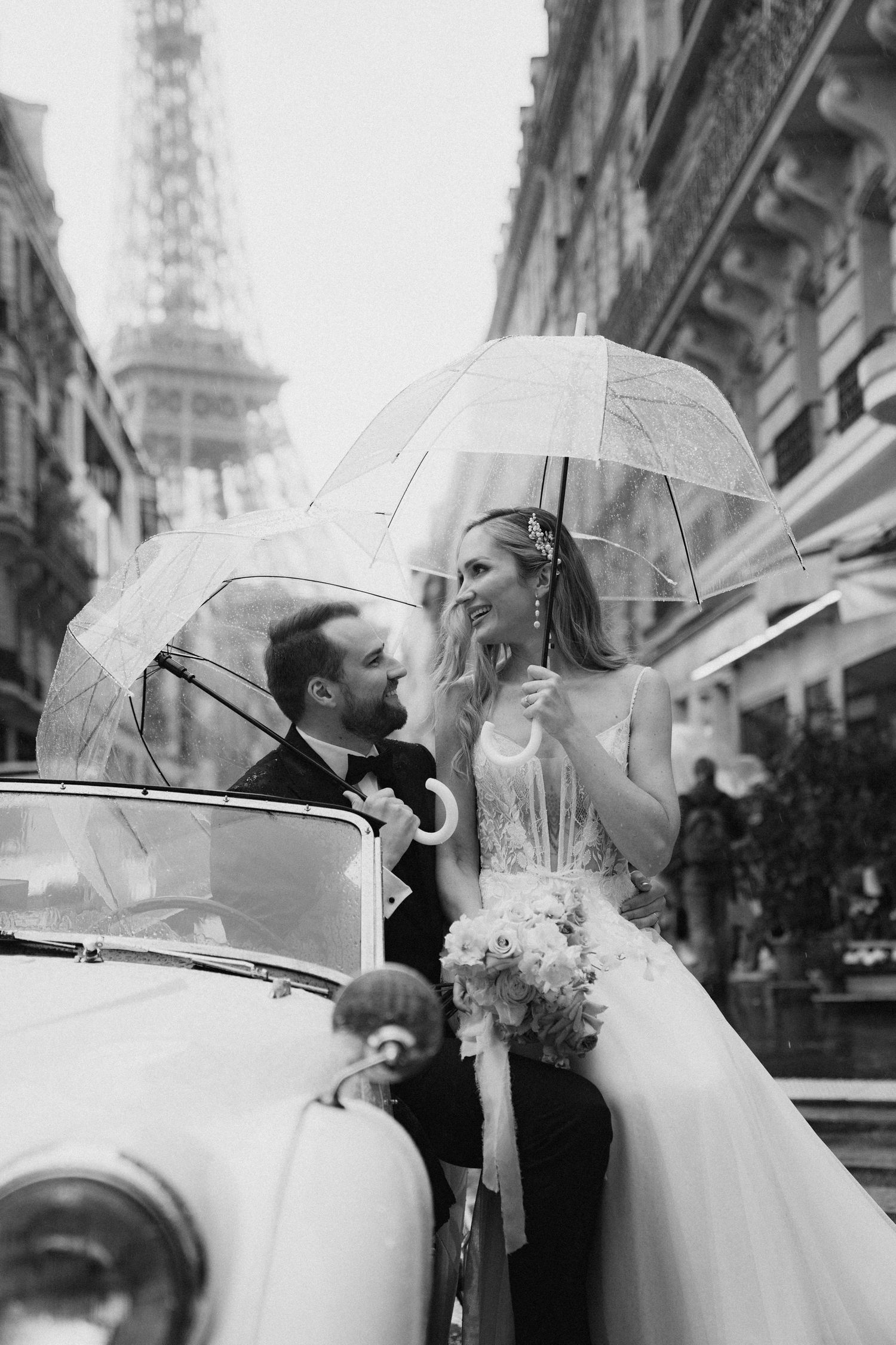 A romantic black and white photo of a couple with clear umbrellas in a vintage car near the Eiffel Tower in Paris.