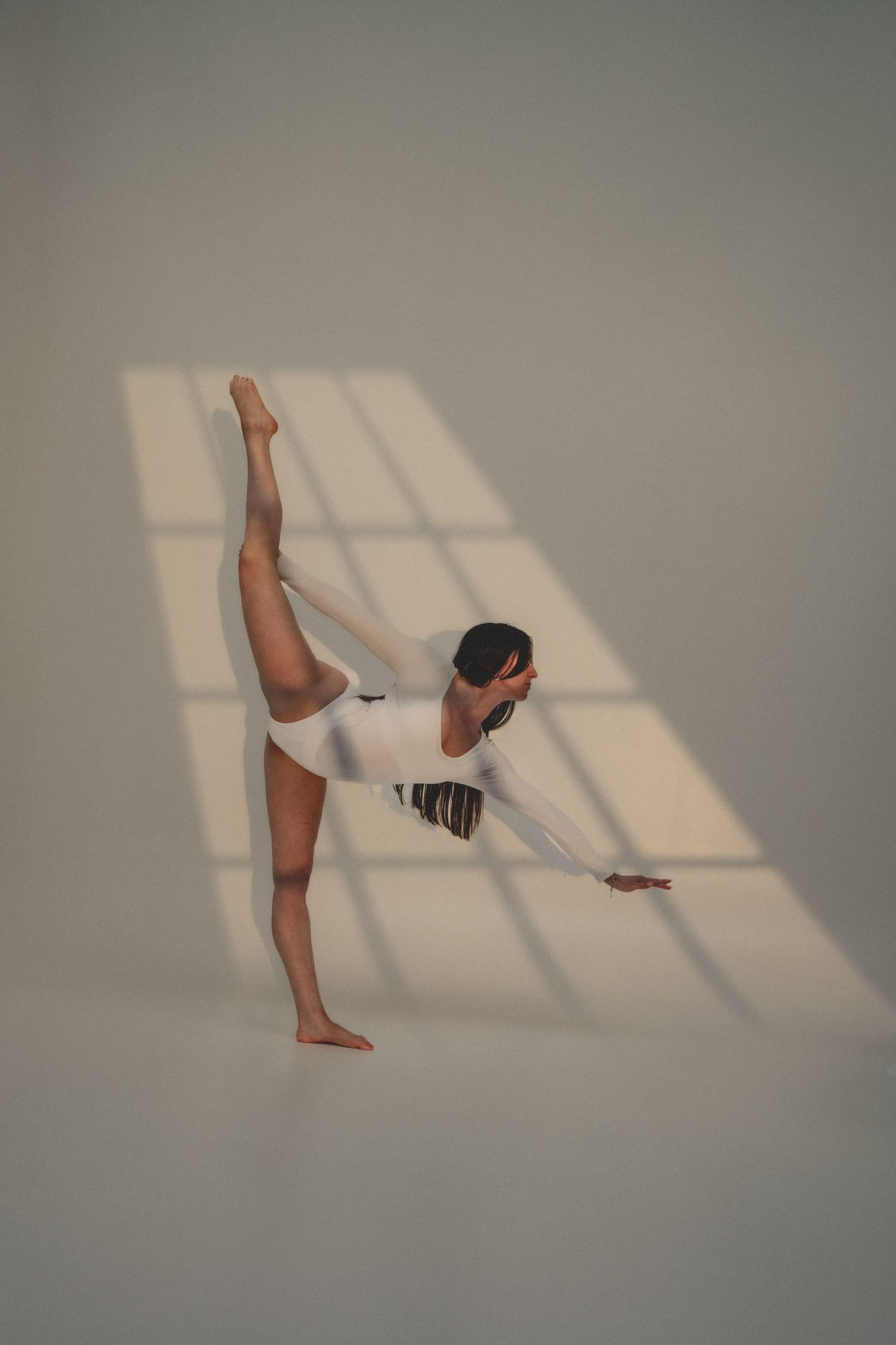 A flexible dancer in white performs a standing split in a sunlit studio, with window light patterns on the wall.
