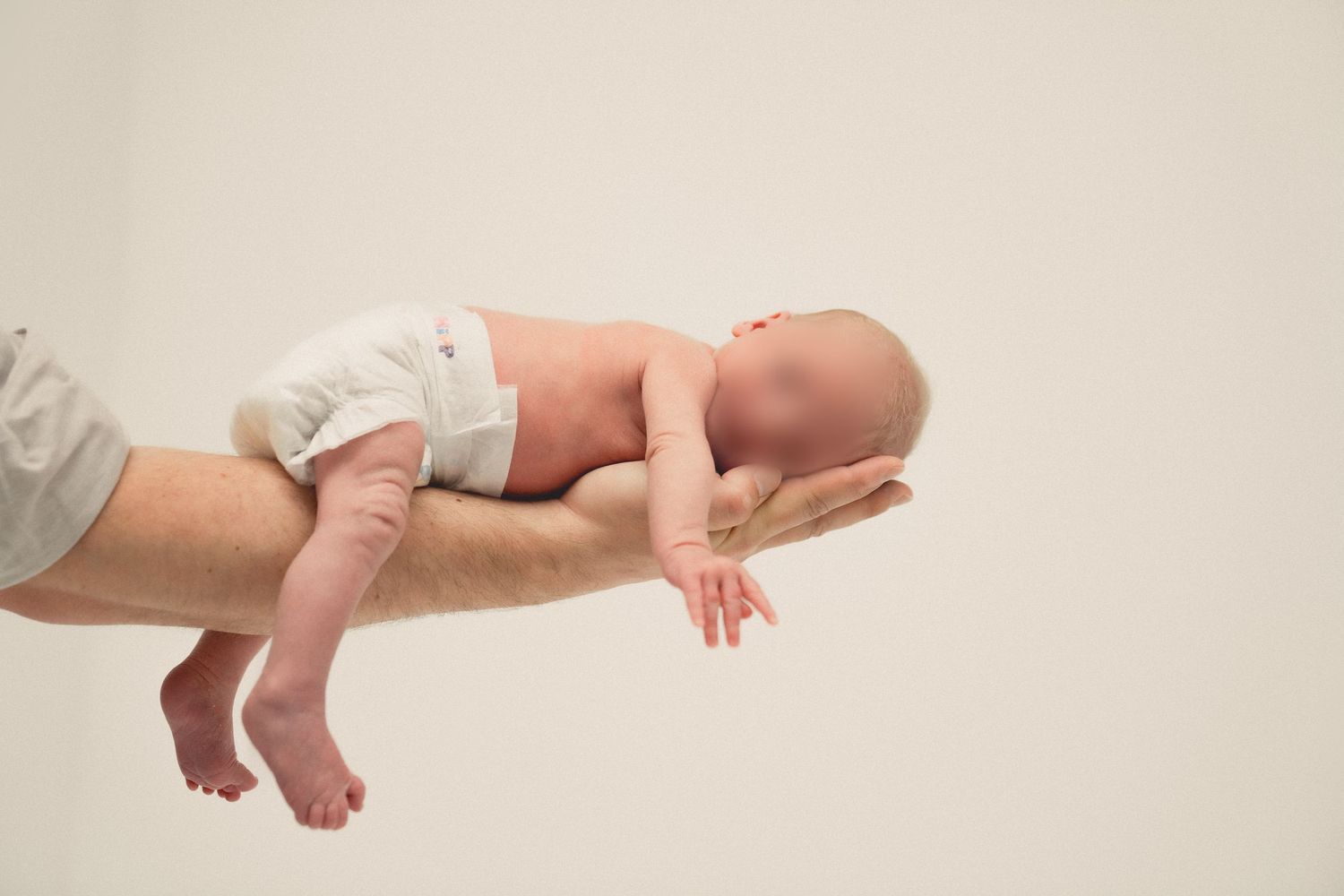 Newborn baby in a white diaper lying on an adult's forearm against a light background.