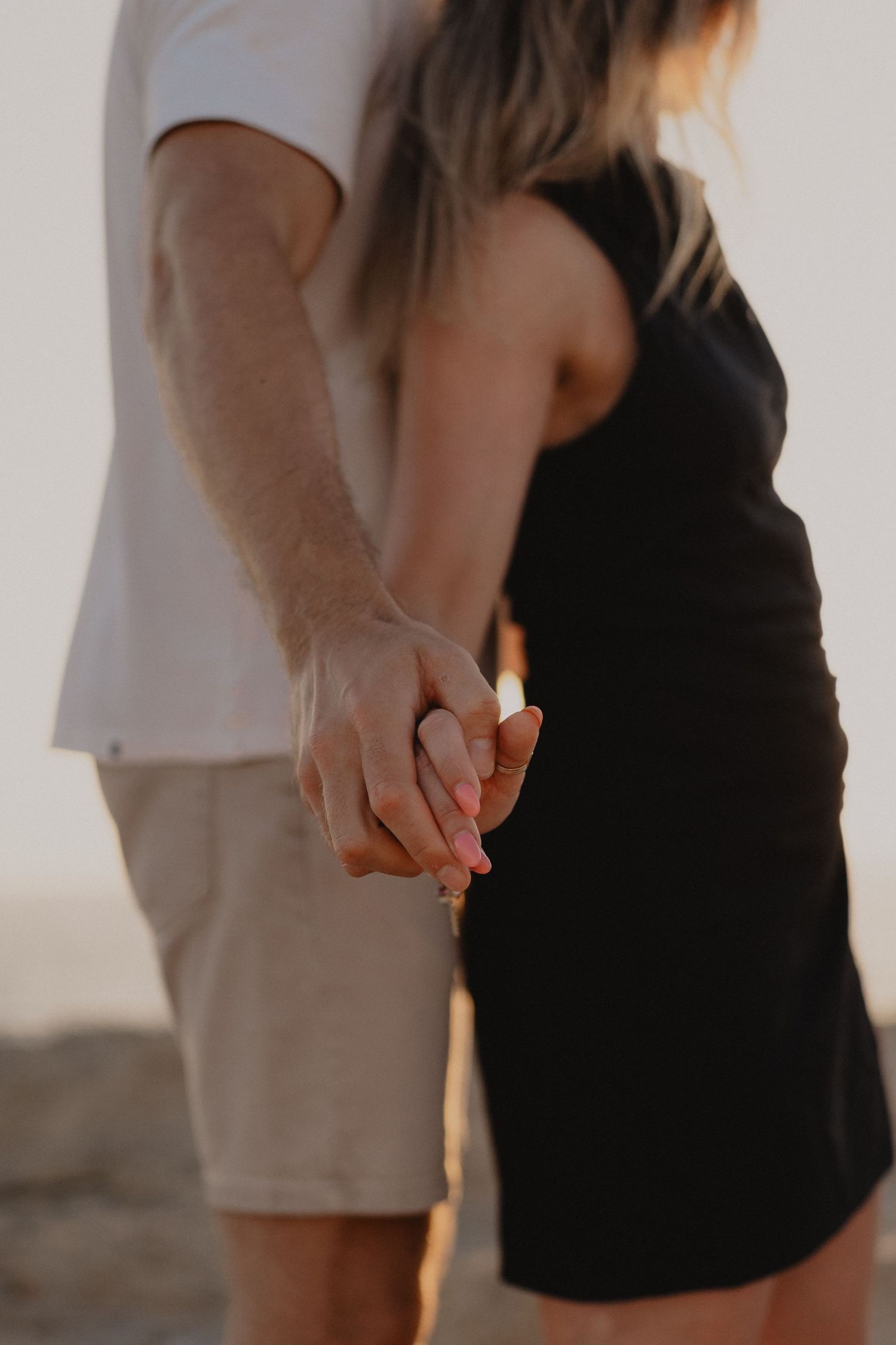 Close-up of a couple holding hands at sunset, woman in black dress and man in white shirt and khaki shorts.