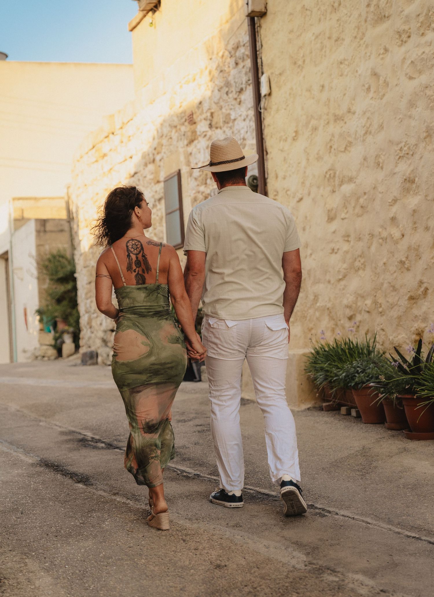 Couple walking hand in hand through a sunny Mediterranean alley, woman in green dress with tattoo, man in white pants and hat.