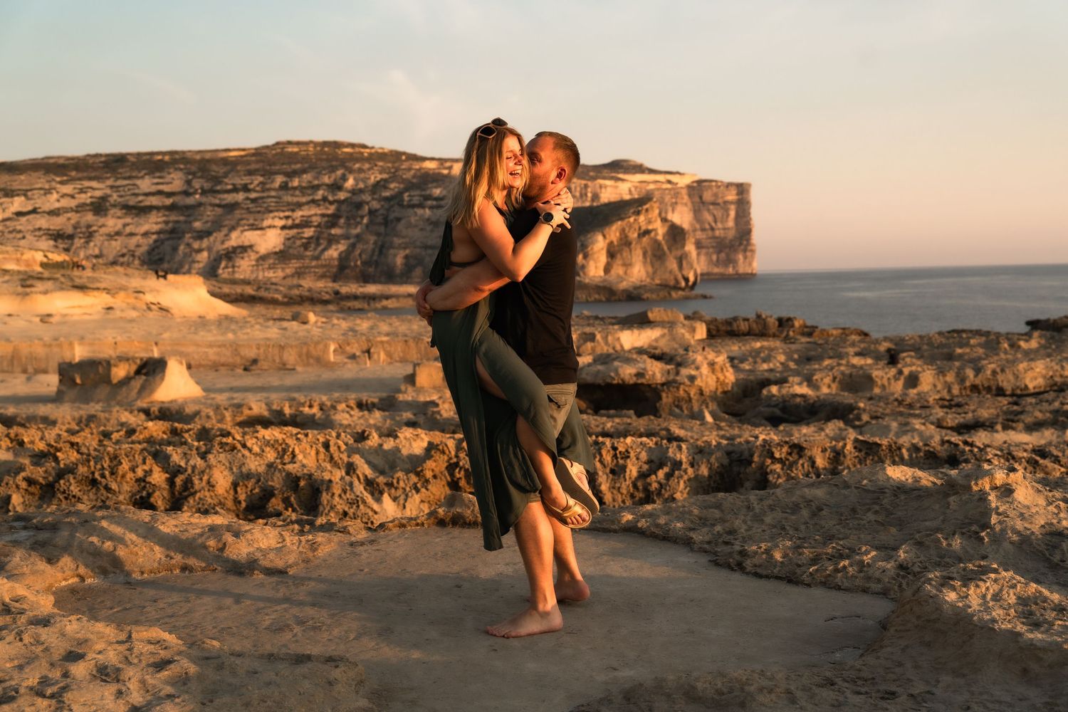 Couple embracing romantically at golden sunset on rocky coastal cliffs by the sea.
