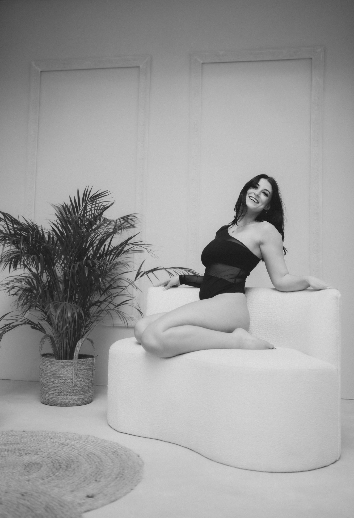 Black and white photo of smiling woman in black bodysuit relaxing on white curved sofa.