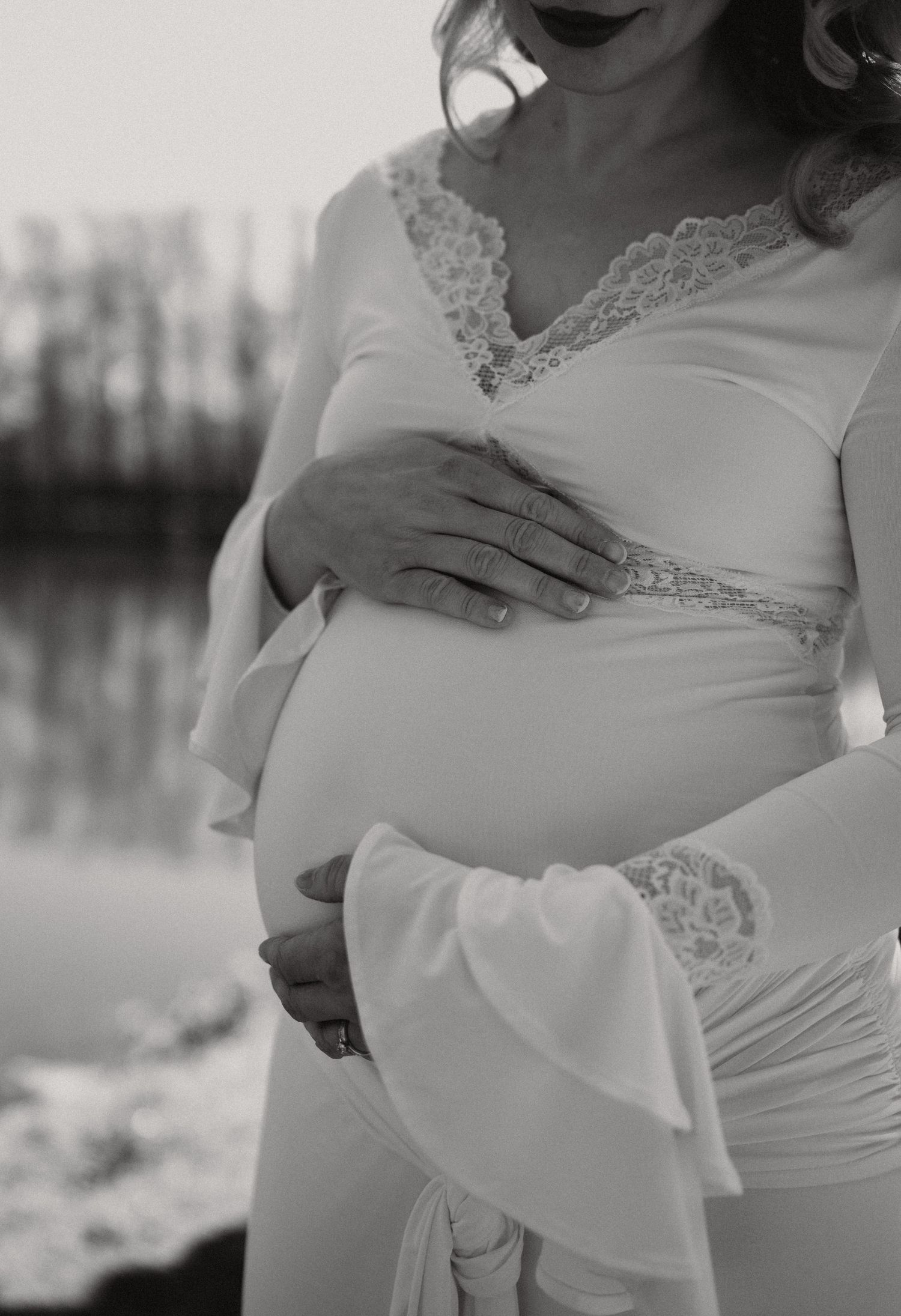 Black and white close-up of a pregnant woman in a white lace maternity dress gently holding her baby bump outdoors.