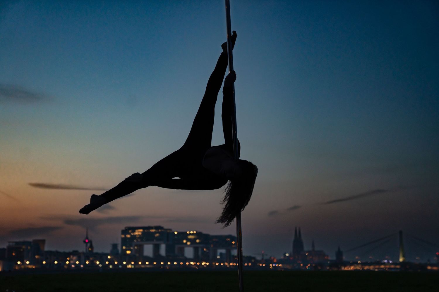 Silhouette of aerial dancer performing on rope at sunset with city skyline in background.