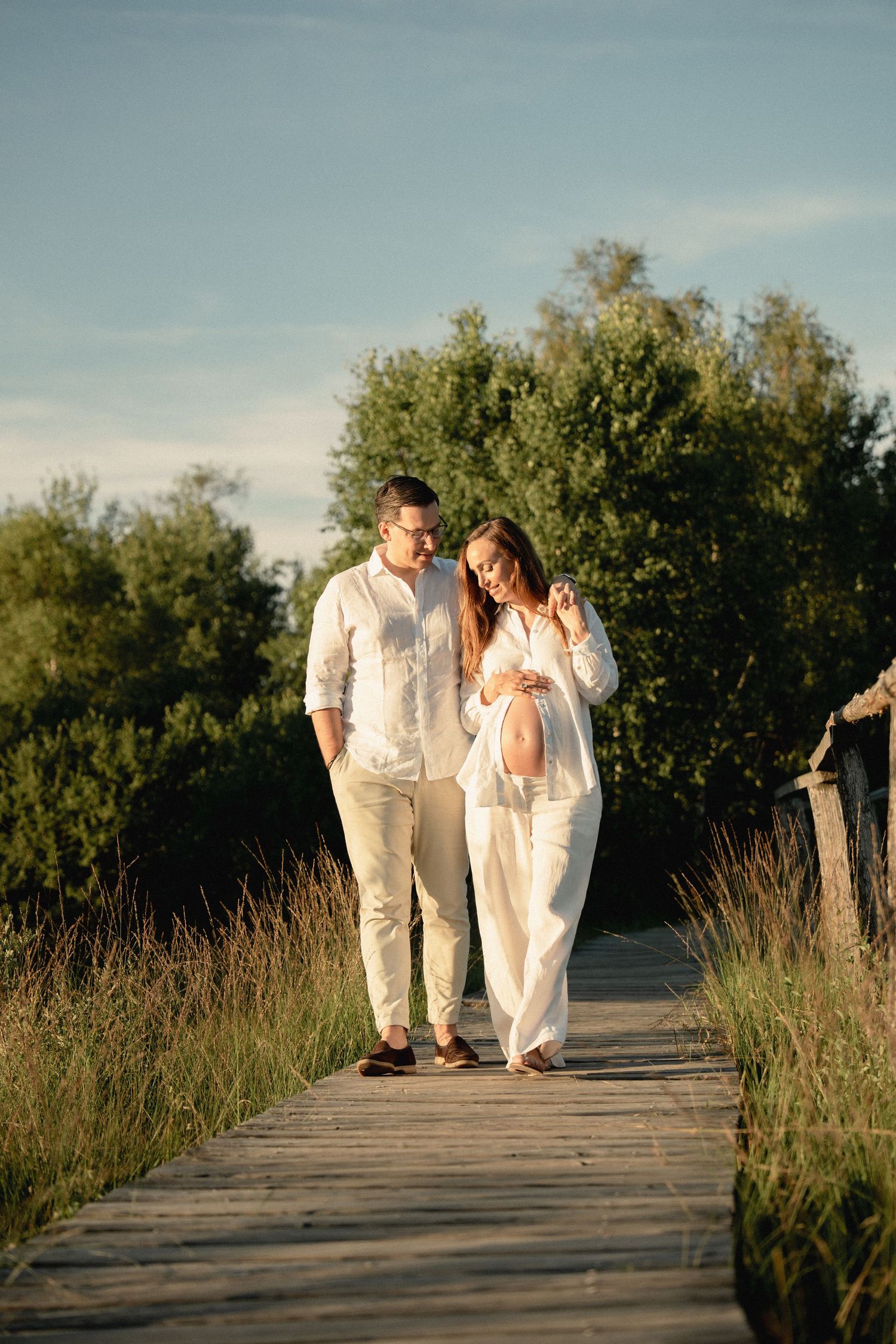 Pregnant couple in white outfits walking on a wooden boardwalk surrounded by green trees in golden hour sunlight.
