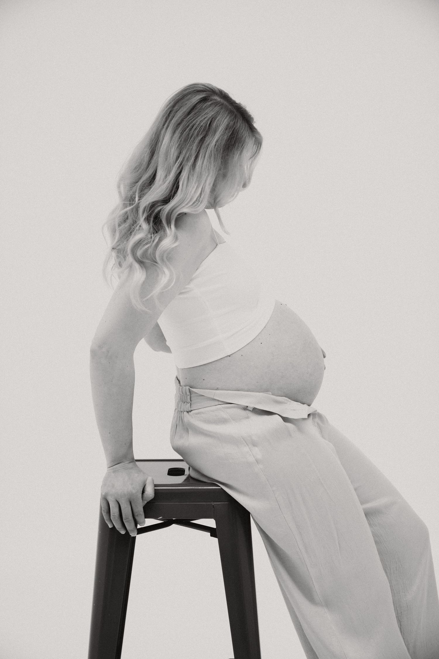 Black and white photo of a pregnant woman sitting on a stool, wearing a crop top and loose pants, showing her baby bump.