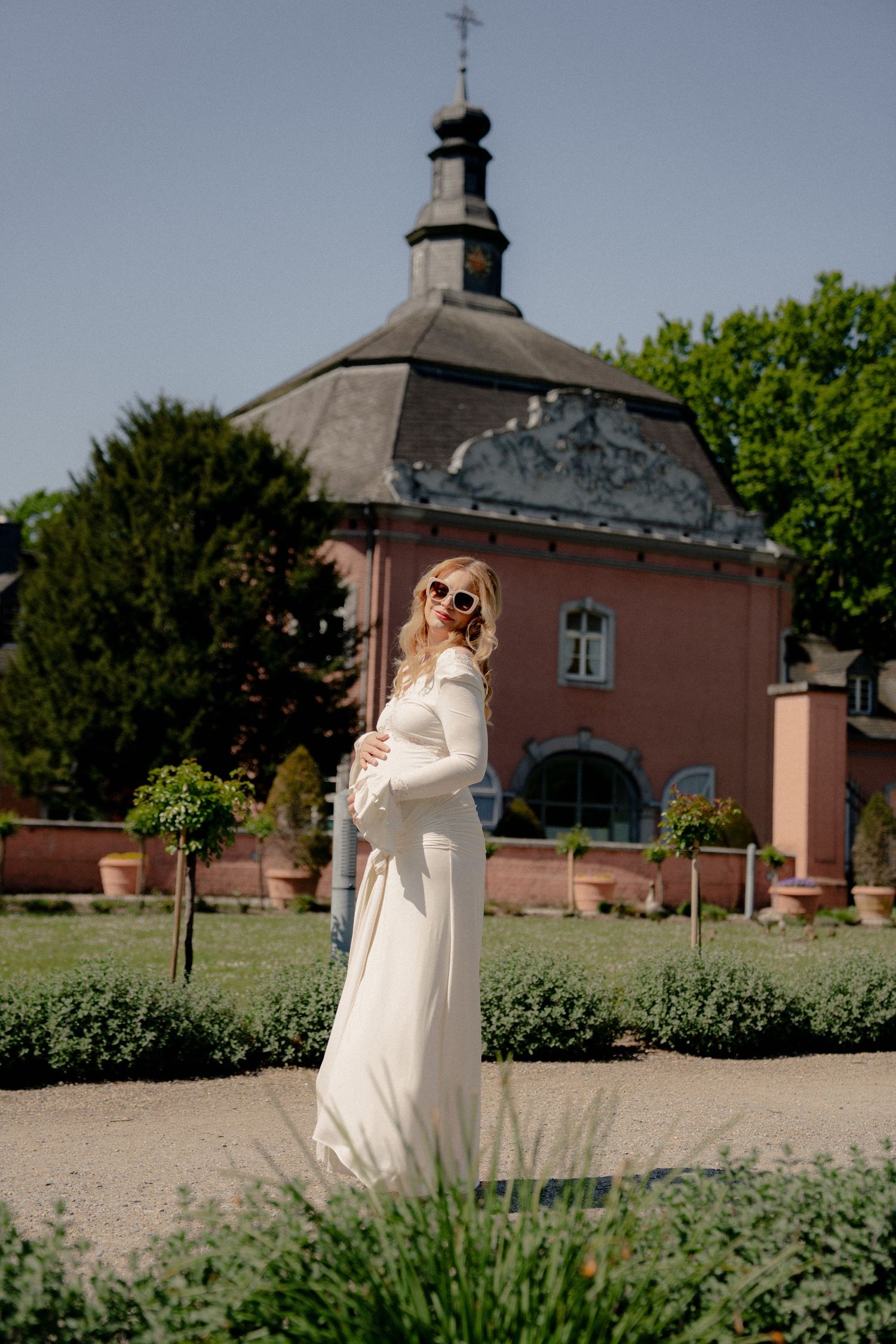 Pregnant woman in white maxi dress poses in front of a pink historic building on a sunny day.